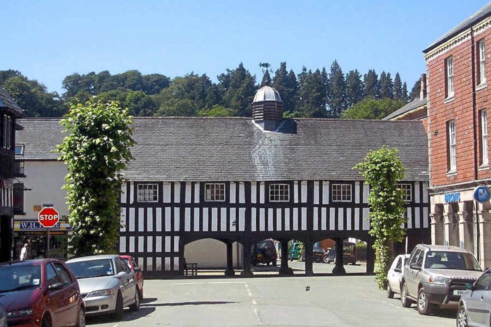 A historic black and white timber-framed market hall spans a street lined with parked cars. Trees flank the building, and a small cupola sits on its roof. A "stop" sign is visible to the left. Forested hills are in the background.