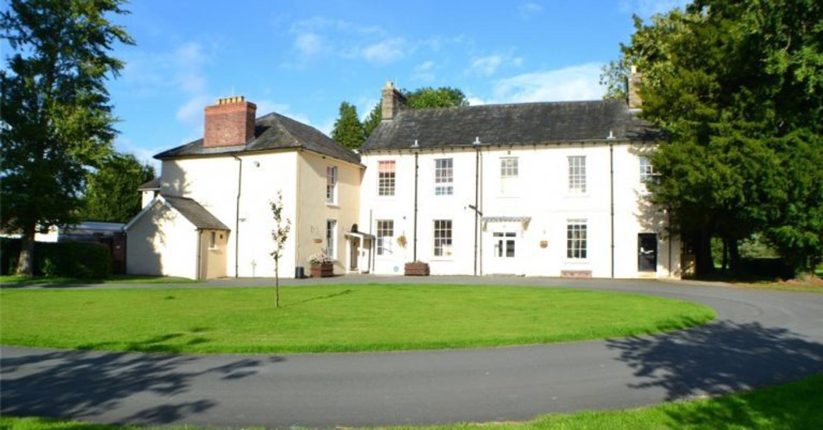 A large, white, two-story house with a dark roof is surrounded by green lawns and trees. A curved driveway leads to the entrance. The sky is clear and blue.