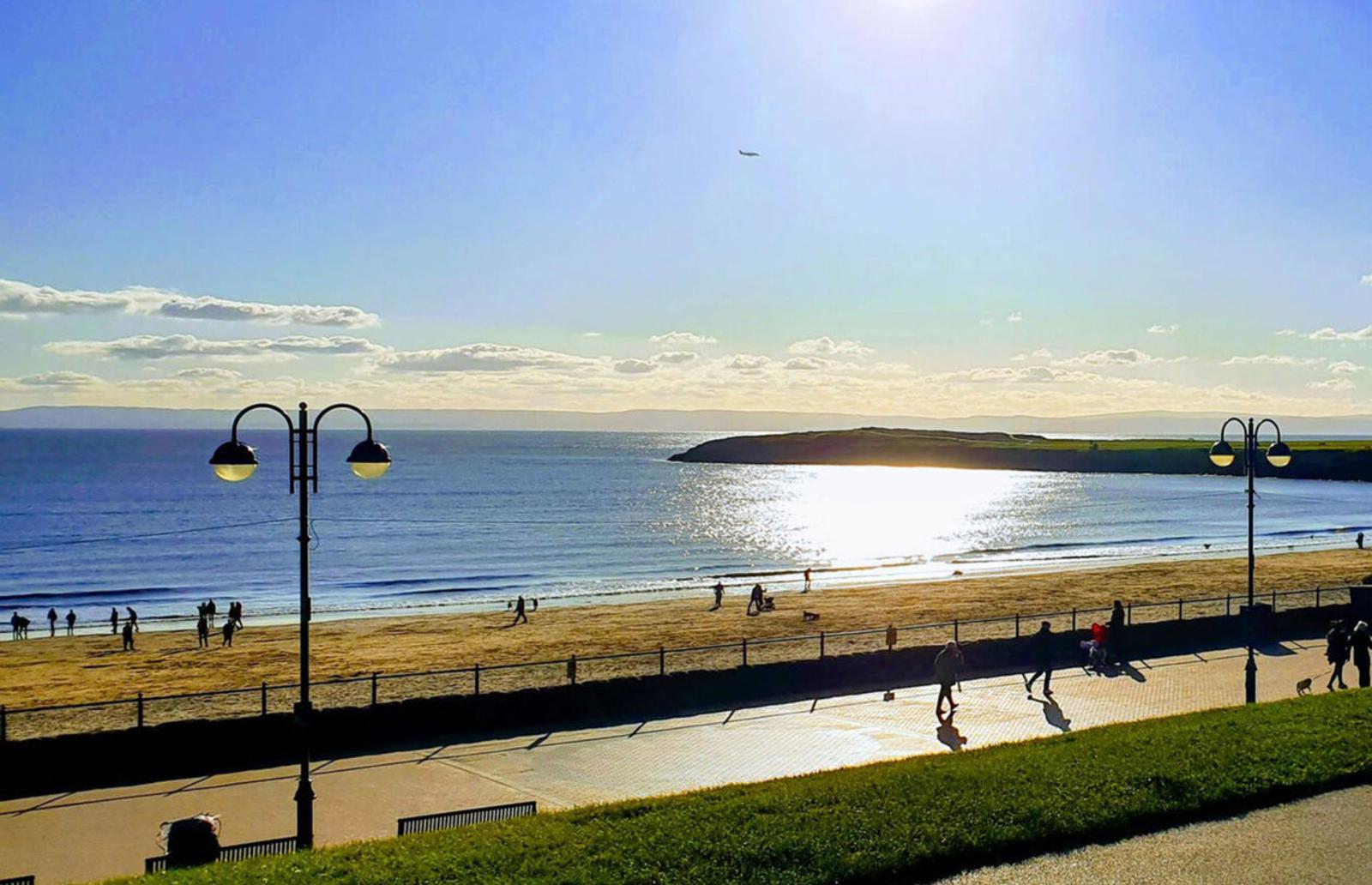 A sunny beachfront scene with a broad beach, scattered people walking and playing, and the sea reflecting sunlight. There is a grassy area and pathway in the foreground, and a hill with a plane in the distant sky.