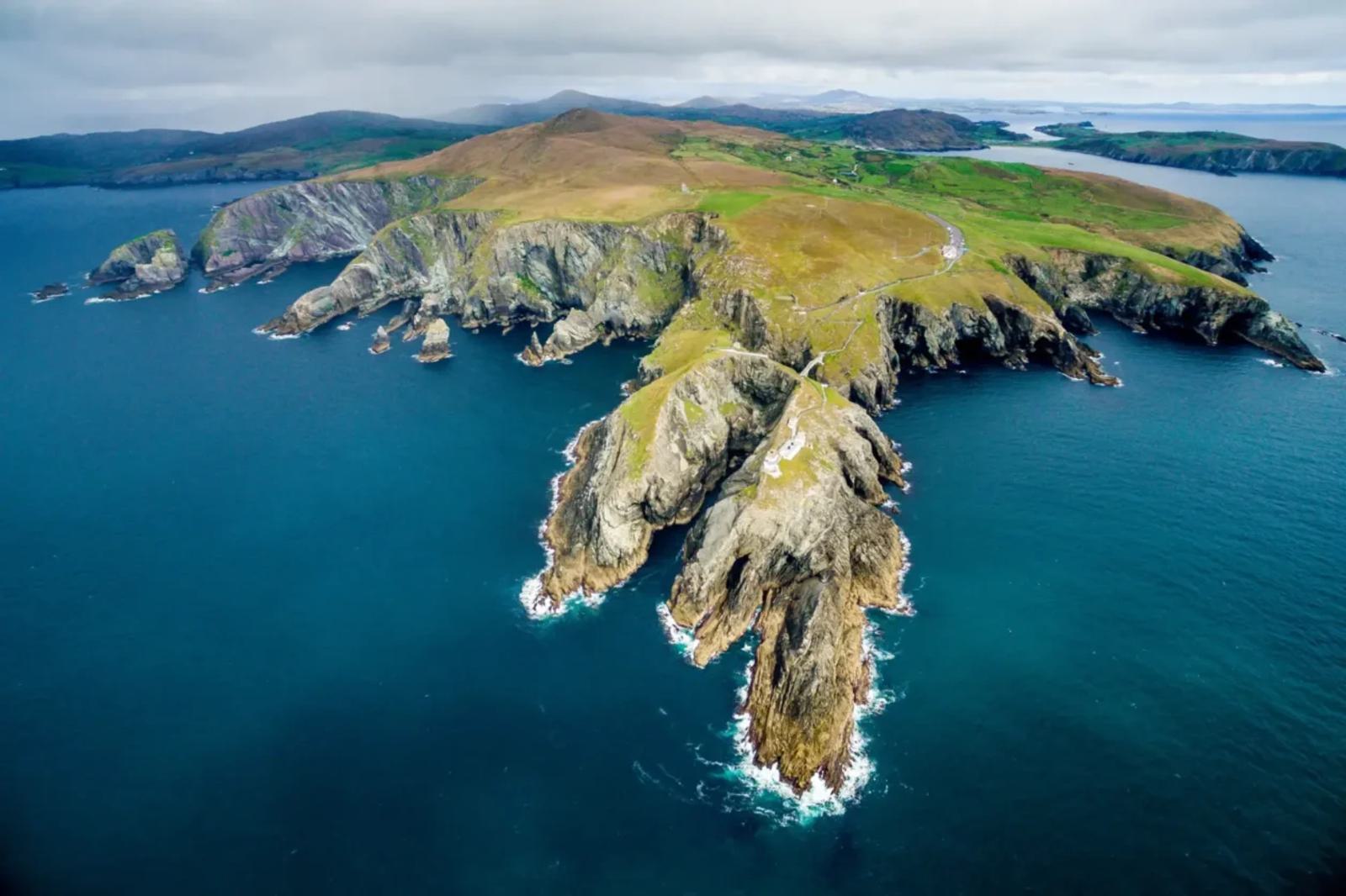 Aerial view of a rugged, rocky coastline with steep cliffs jutting into the ocean. The landscape is covered in patches of green grass and surrounded by deep blue water. Cloudy sky in the background adds a dramatic tone to the scene.