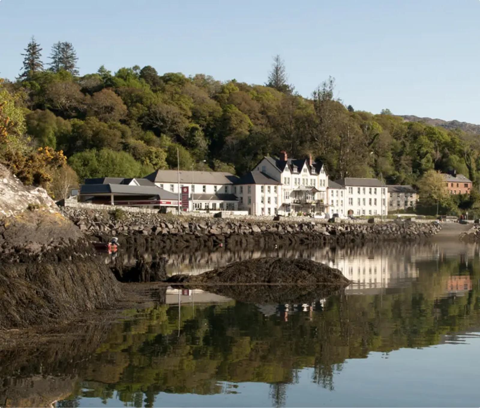 A scenic view of a small town with white buildings lining a rocky shoreline. The structures are reflected in the calm water, bordered by lush green trees on a hillside under a clear blue sky.