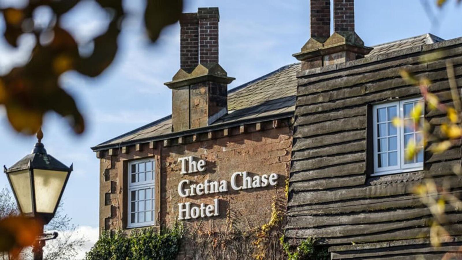 A two-story brick building with ivy, featuring a sign that reads "The Gretna Chase Hotel." Two chimneys are visible on the roof, and a classic streetlamp stands nearby. Leaves frame the image in the foreground.