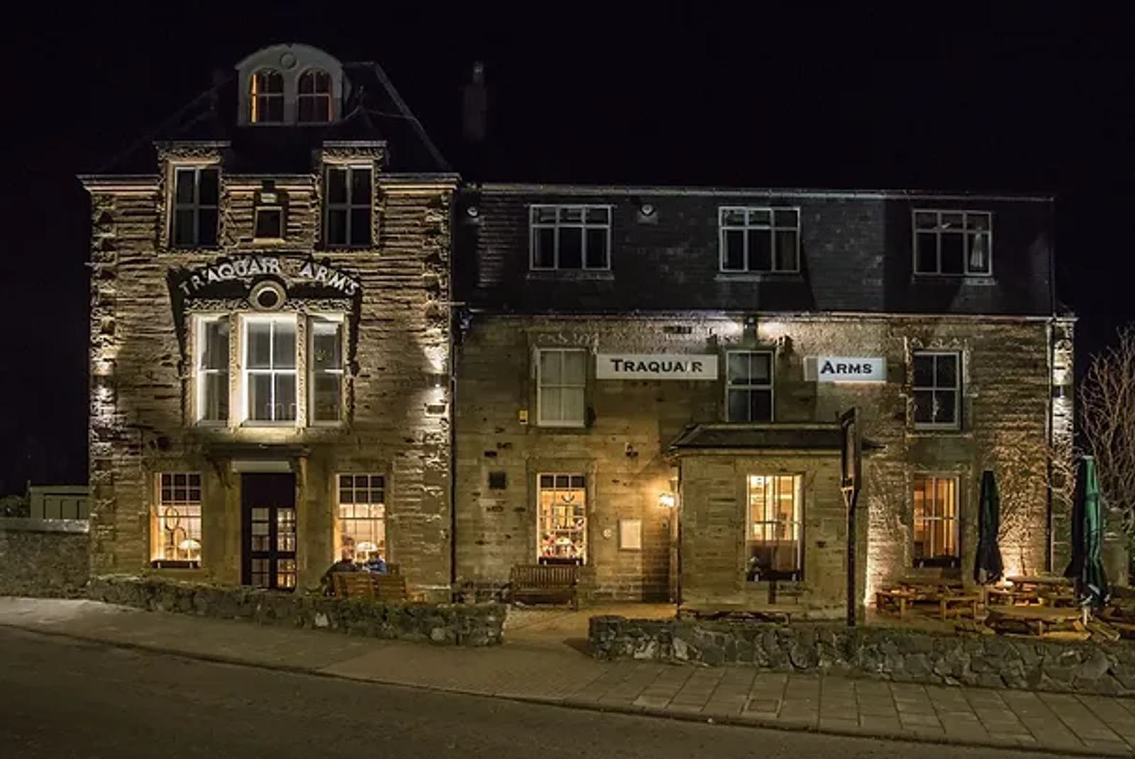 A large, three-story stone building, the Traquair Arms, is lit up at night. Its sign glows warmly above the entrance. Wooden benches and tables sit outside on the right. The street in front is empty.