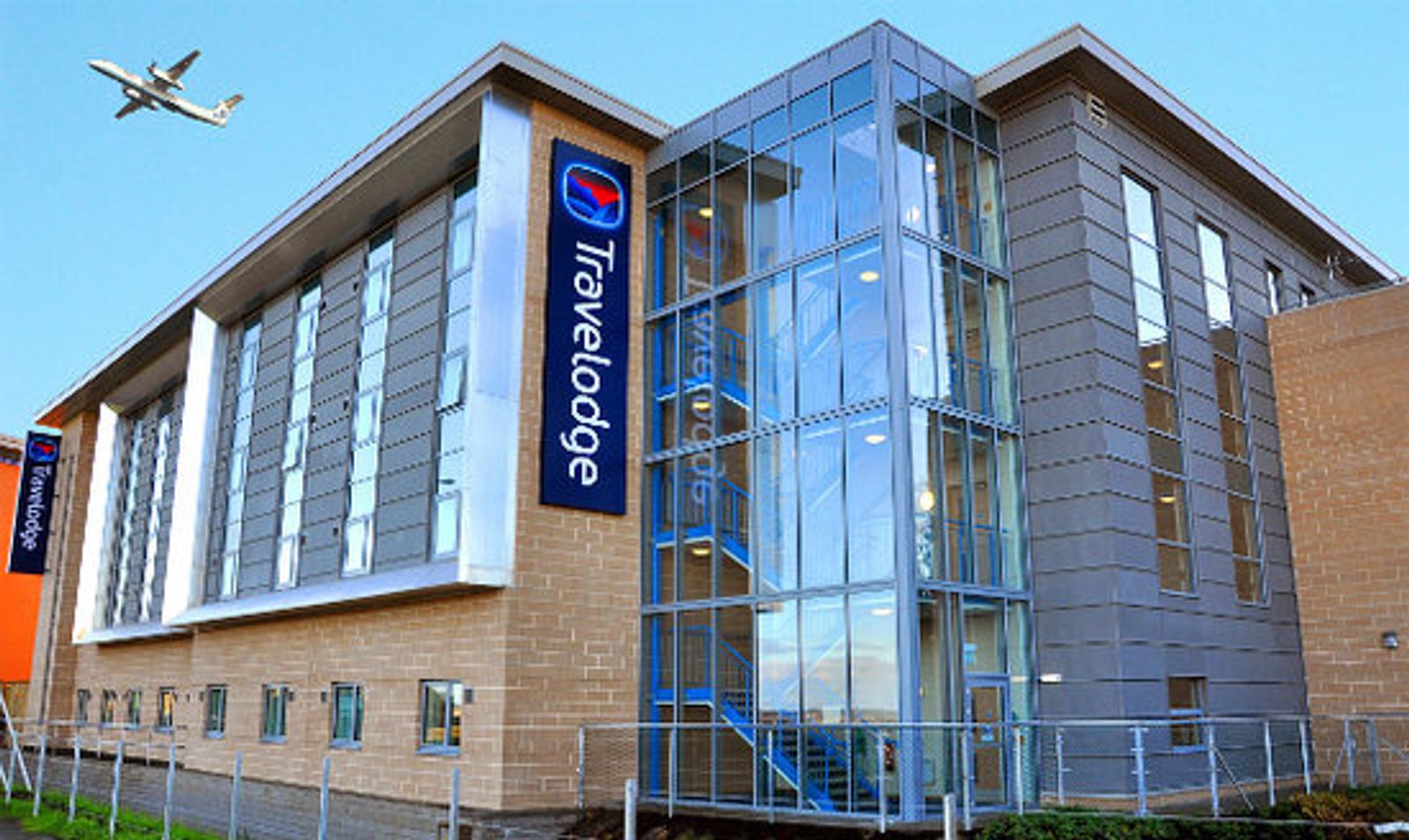 A modern glass and brick building with a Travelodge sign. Large windows and a metal fence are visible. There's an airplane flying in the sky above.
