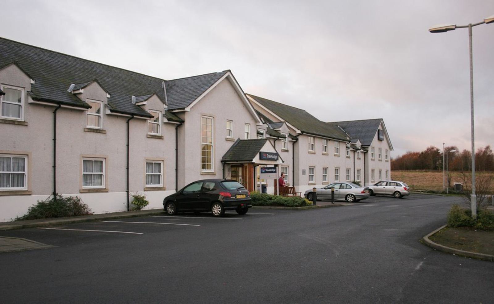 A two-story white building with a sloped roof and multiple windows, resembling a hotel or inn. Several cars are parked in the paved lot in front. The sky is overcast, and there is a grassy field in the background.