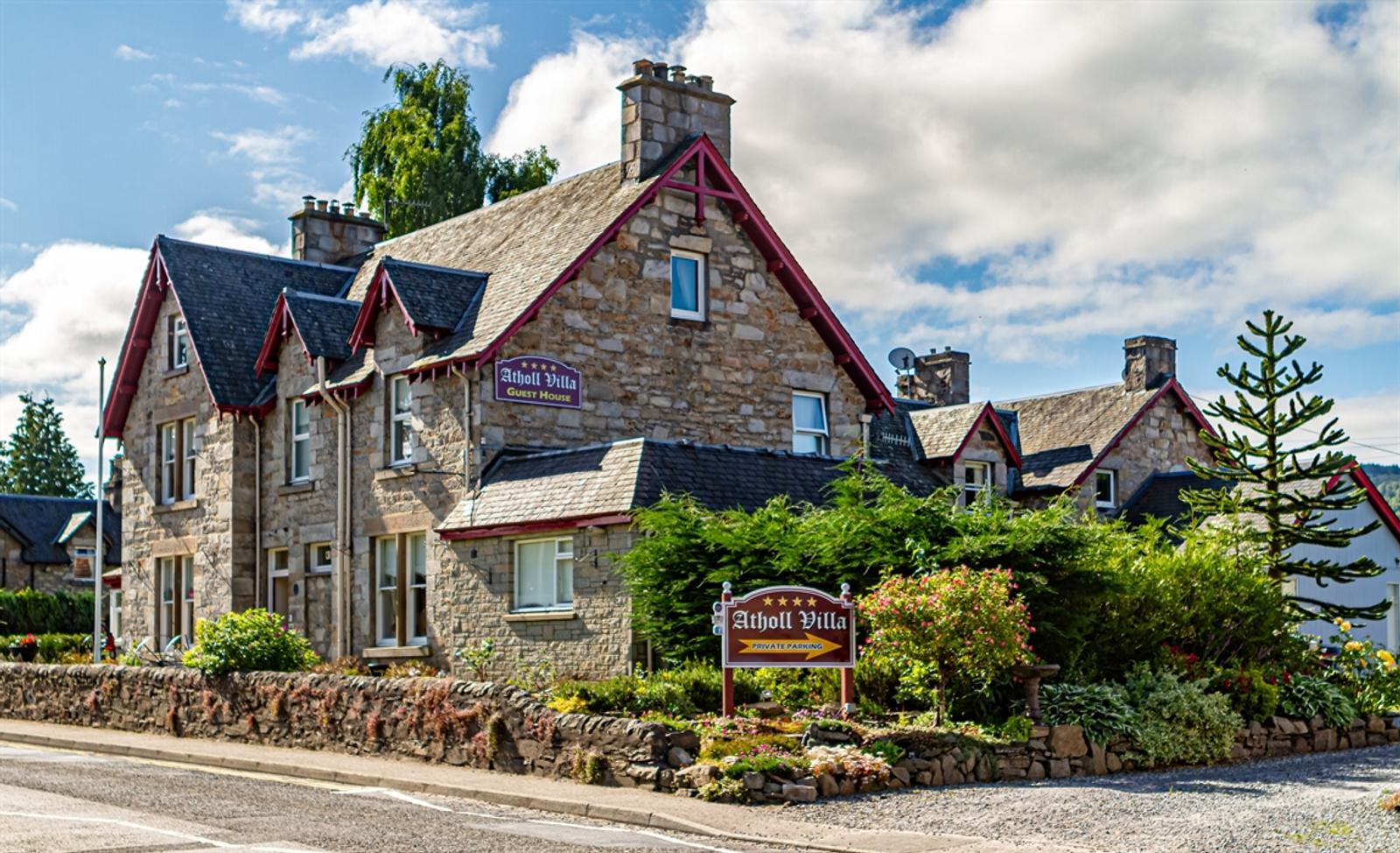 A charming stone guest house with multiple peaked roofs, red trim, and chimneys, surrounded by lush greenery and colorful flower beds. A sign reads "Atholl Villa Guest House." The sky is partly cloudy, enhancing the inviting atmosphere.
