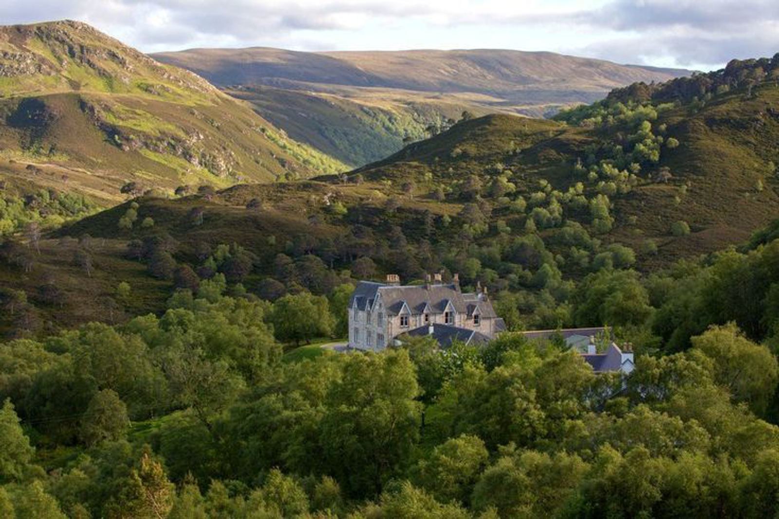 A large stone building nestled among lush green trees, set in a valley surrounded by rolling hills and mountains. The sky is partly cloudy, casting soft light over the landscape.