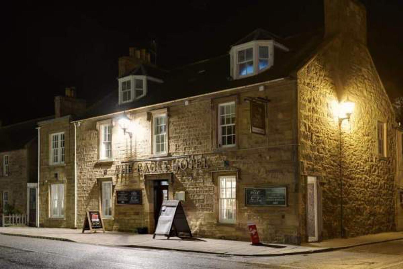 Nighttime view of a stone building with a sign reading "The Eagle Hotel." Warm lights illuminate the facade, displaying chalkboard signs near the entrance. The street in front is empty, and the scene is quiet and inviting.