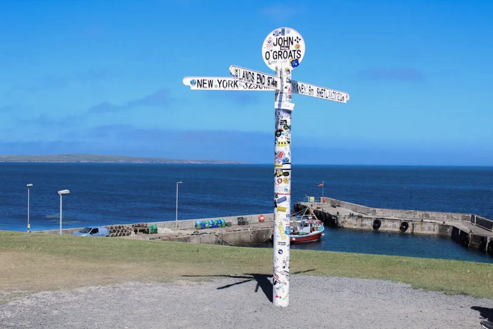 Signpost at John o' Groats, Scotland, with directional signs pointing to New York and other locations, covered in stickers. A small harbor and calm blue sea are visible in the background under a clear blue sky.