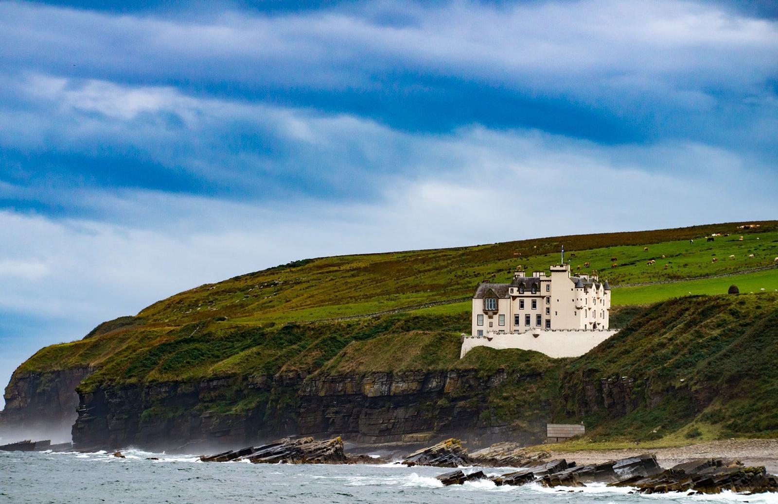 A white castle stands atop a lush green cliff overlooking a rocky coastline with gentle waves. The sky is overcast with patches of blue, creating a serene and picturesque landscape.
