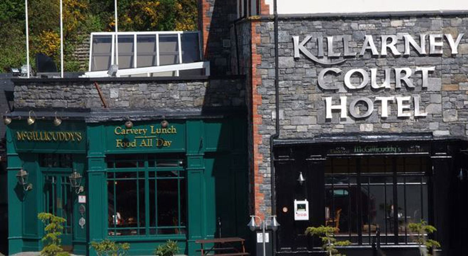 Exterior of Killarney Court Hotel with stone facade. To the left, McGillycuddy's pub features green paint and signage for "Carvery Lunch Food All Day." Both establishments have traditional architectural details.