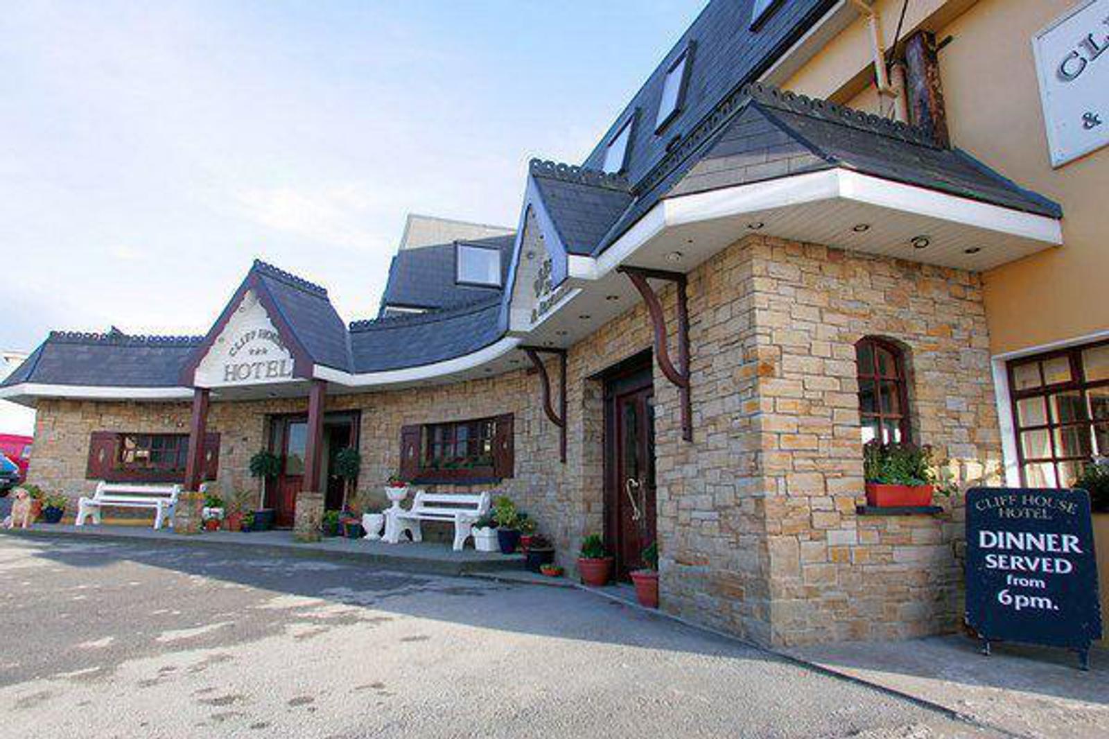 A stone-fronted hotel with decorative windows and a pitched roof. There are white benches and potted plants outside. A sign announces dinner service starting at 6 PM. The sky is clear.