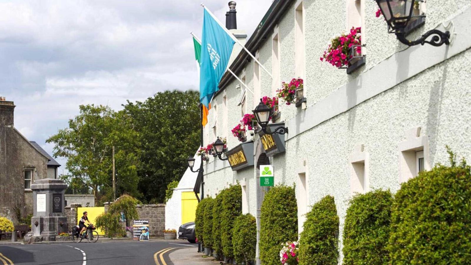 A quaint stone building with hanging flower baskets has flags on the roof. A cyclist rides past and a pedestrian walks near a monument on a narrow road lined with bushes. The sky is partly cloudy.