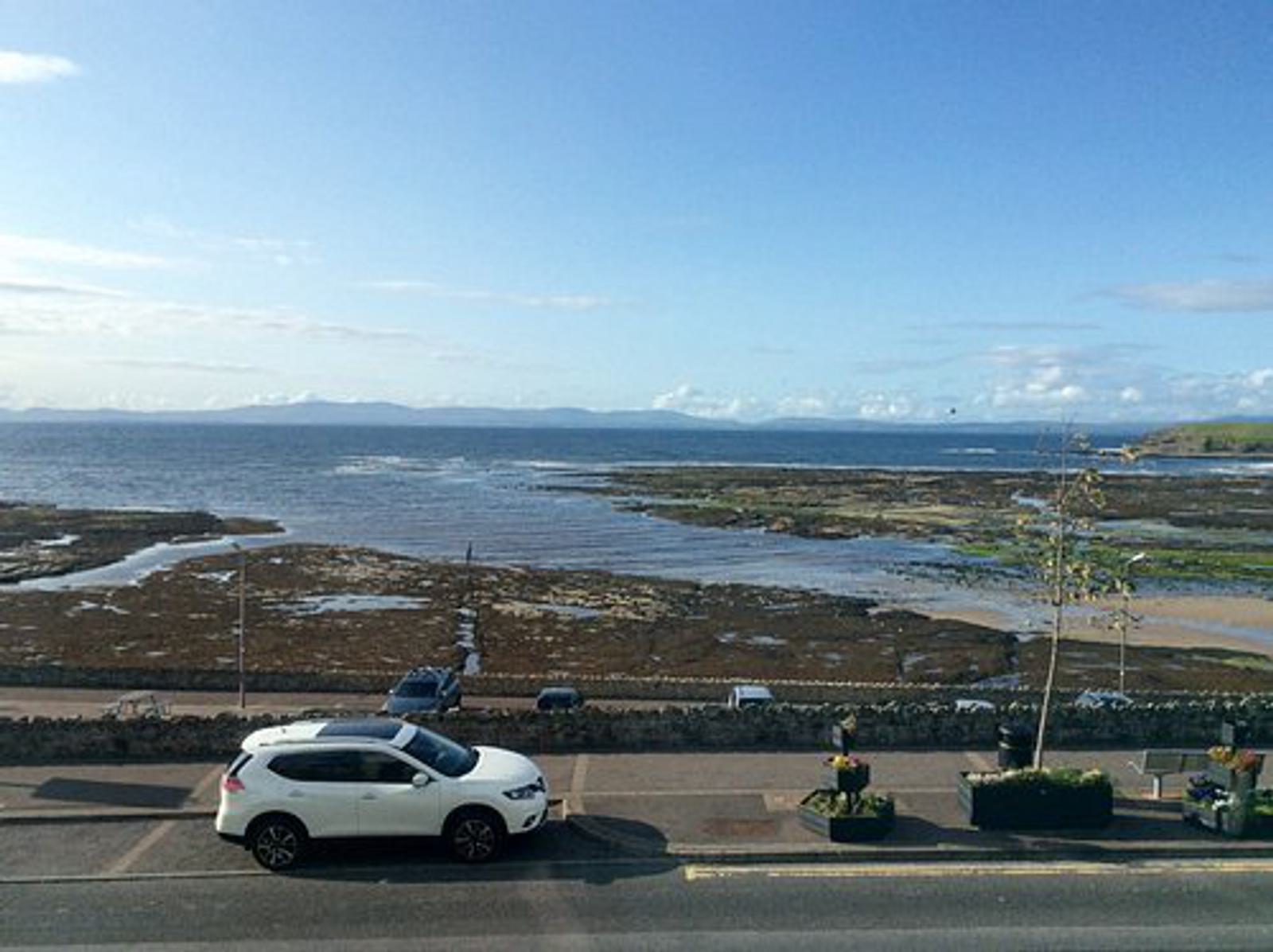 A coastal scene featuring a white car parked alongside a road with a stone wall. Beyond the wall is a rocky shoreline with pools of water, and a vast expanse of blue sea stretching to the horizon under a clear blue sky.