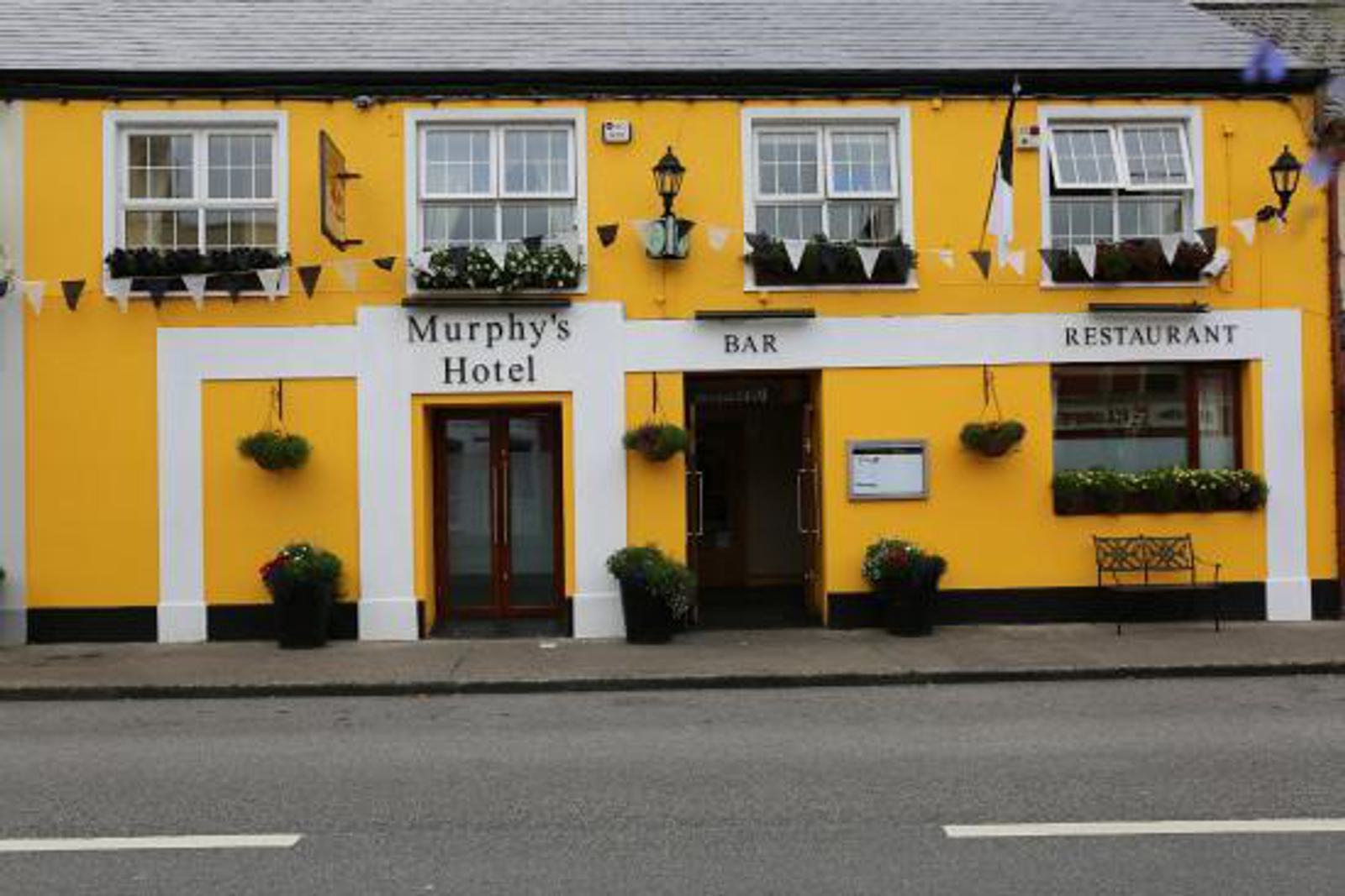Yellow building with white trim labeled "Murphy's Hotel" featuring upper windows with flower boxes. Signs indicate a bar and restaurant. There are potted plants flanking the entrance, and a bench is placed outside on the pavement.