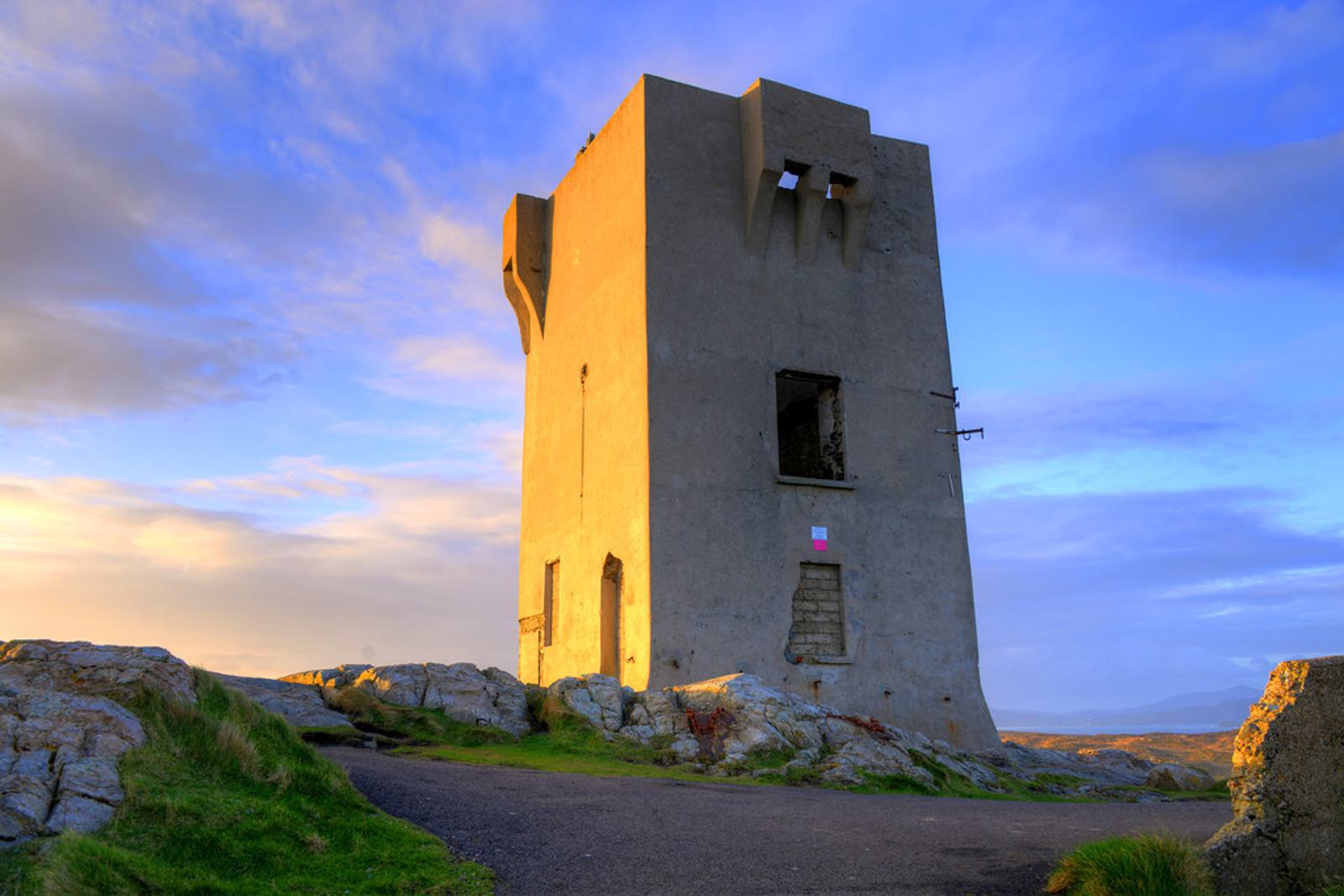 A tall, weathered stone tower stands on a rocky landscape under a blue sky with scattered clouds. The sun casts a golden glow on the structure, highlighting its rugged texture and angular shape. Grass and rocks surround the base.