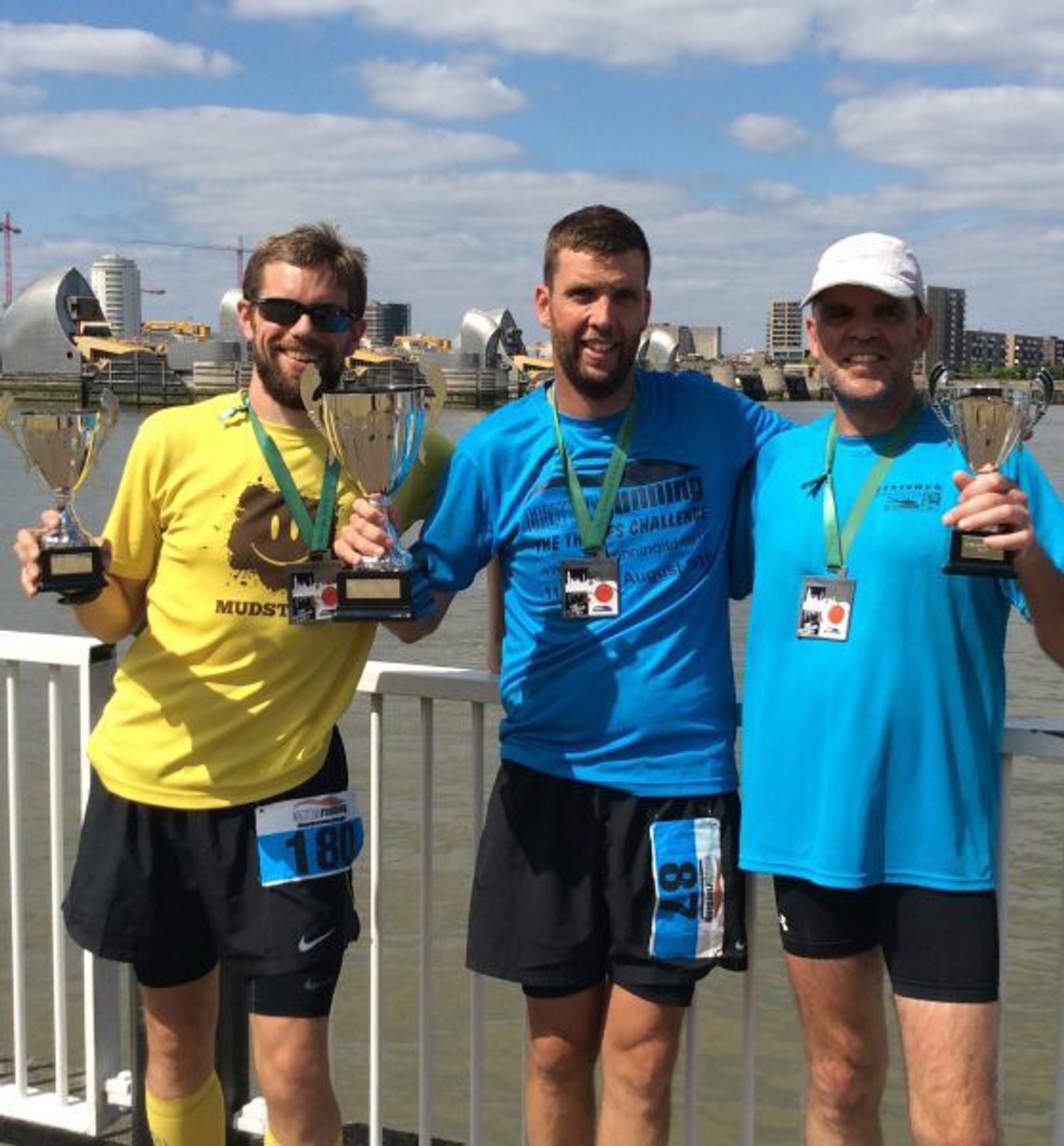 Three men stand outdoors on a sunny day, holding trophies and wearing medals around their necks. They are dressed in running attire, wearing shorts and t-shirts, and are smiling for the camera. Buildings are visible in the background across a body of water.