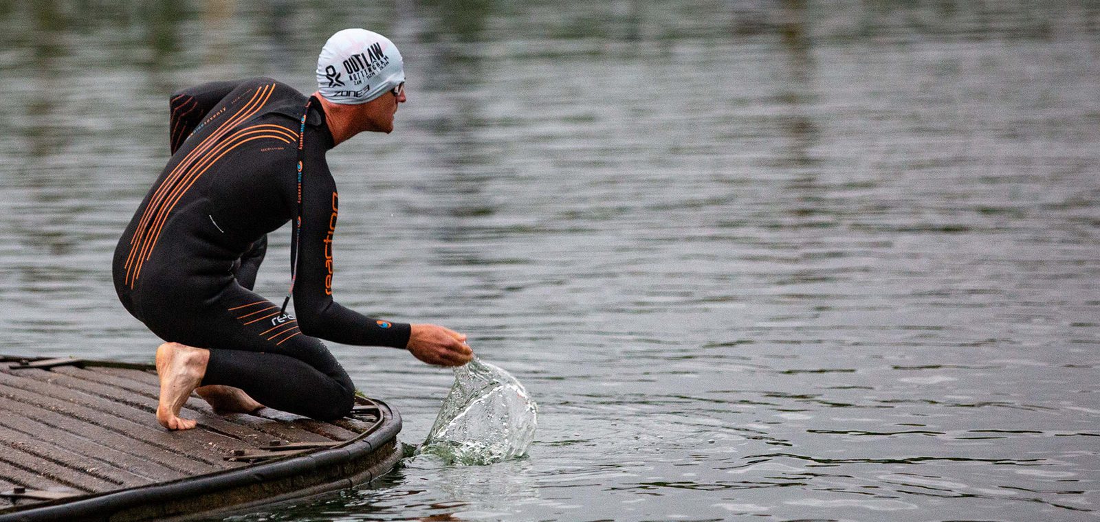 A person in a wetsuit and swim cap stands on a dock while crouching down and touching the water with one hand, causing a splash. The individual appears to be preparing to enter a body of water, possibly for swimming or a triathlon.