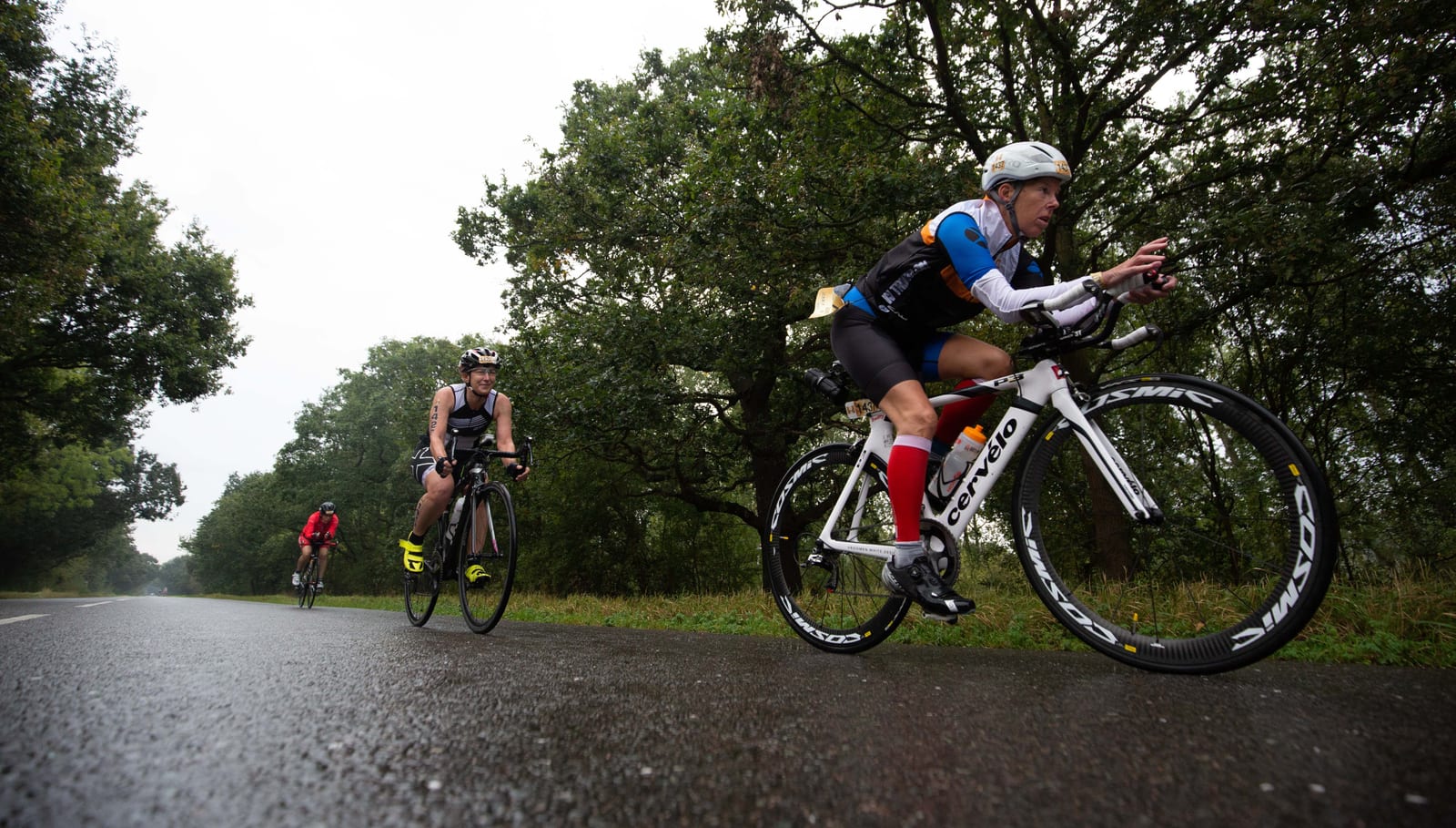 A group of cyclists ride on a wet, tree-lined road. The cyclist in the front wears a white helmet, blue and white jersey, and rides a white bike. Two other cyclists follow, one wearing a red outfit. The scene is set in a lush, green, outdoor environment.