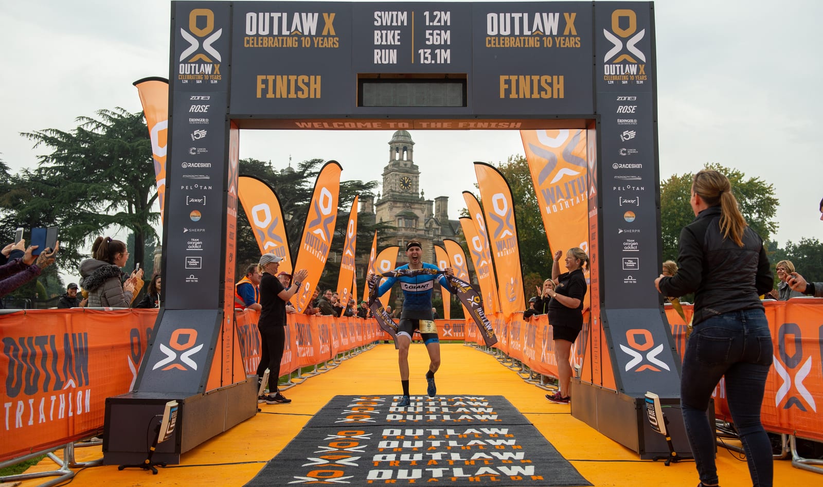 A triathlete crosses the finish line at the Outlaw X Triathlon. The athlete is cheered on by spectators as they run on an orange carpet towards the finish line arch. The event branding and sponsor logos are visible on banners and flags, with a historic building in the background.