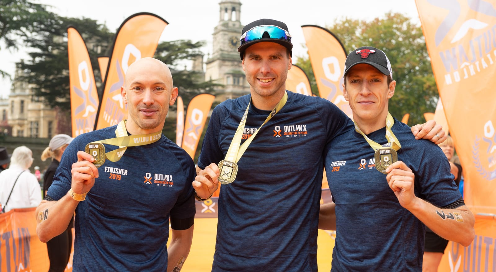 Three men, all wearing blue "Outlaw" t-shirts and displaying their medals, smile at the camera. They are standing outdoors, with orange "Outlaw" banners in the background. The central figure has his arms around the other two.