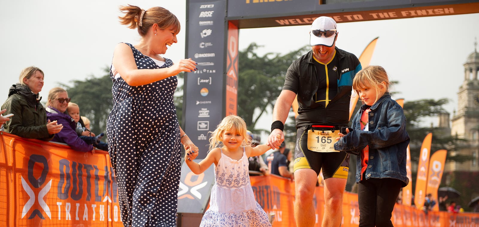 A man wearing athletic gear and a medal crosses the finish line of a triathlon holding hands with two young girls. A woman in a polka dot dress holds one girl's hand, smiling. Spectators stand behind orange barriers, and "Outlaw Triathlon" banners are visible.