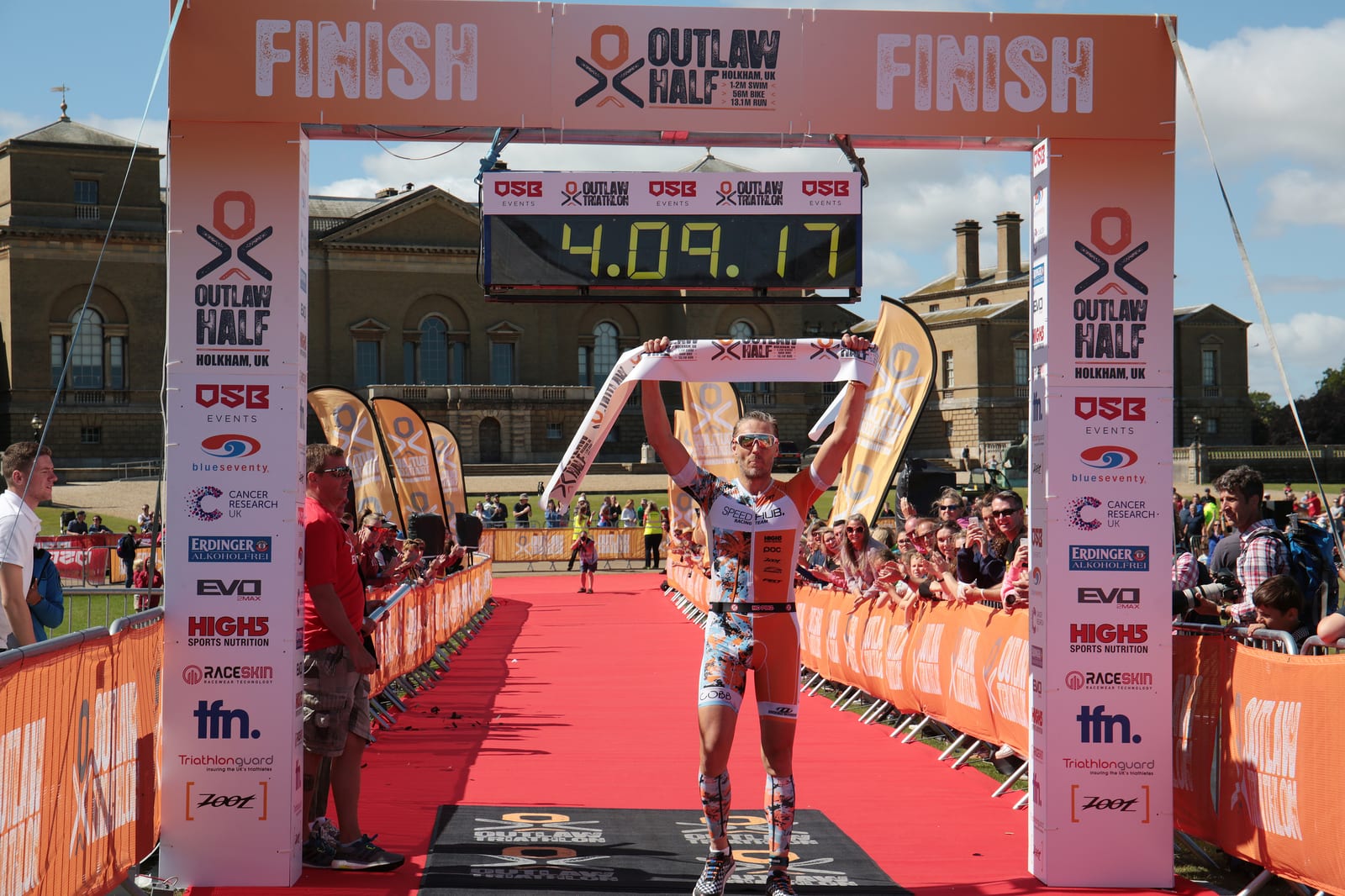 A person in athletic gear raises their arms in victory as they cross the finish line of the Outlaw Half race. The digital timer overhead displays a time of 4:09:17. Spectators line the sides of the finish area, with a historical building in the background.