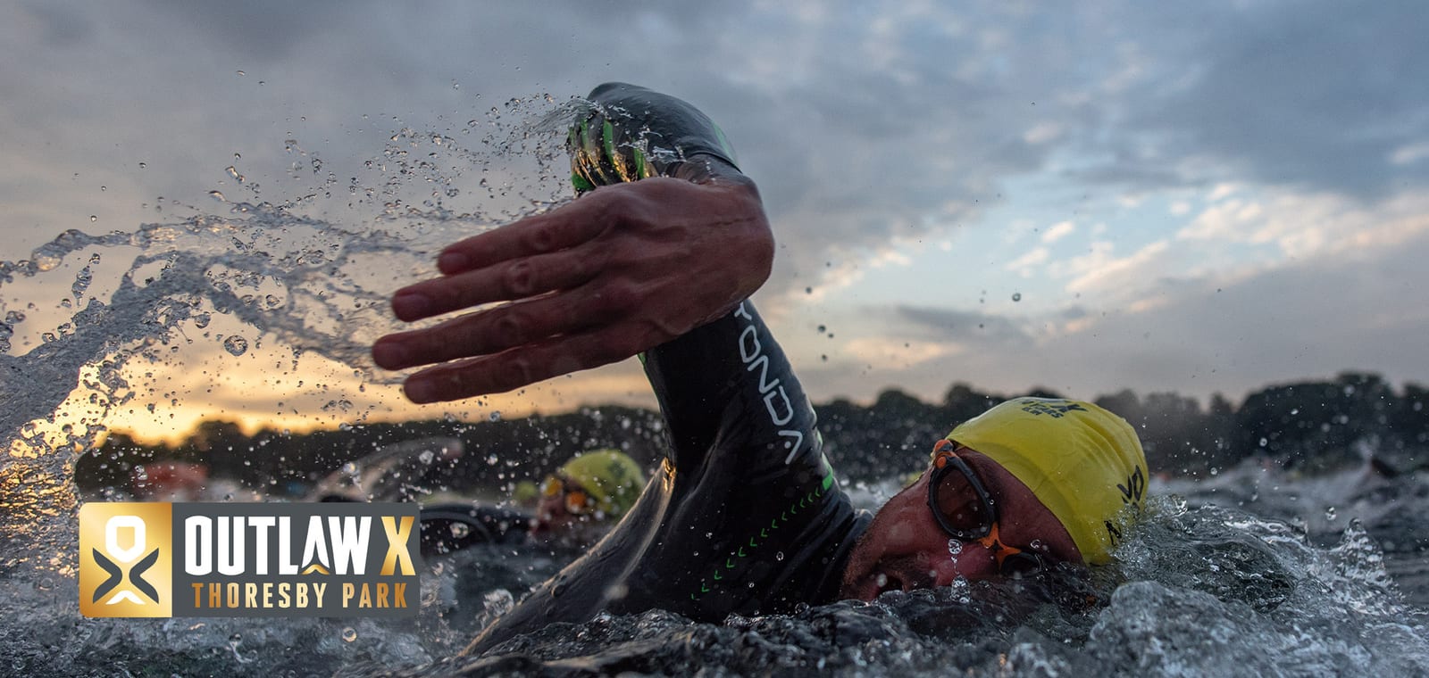 A close-up shot of a swimmer wearing a yellow swim cap and goggles, making a stroke in open water. Other swimmers are seen in the background. The sky is partly cloudy with the sun setting. A logo on the bottom left reads "OUTLAW X, Thoresby Park.
