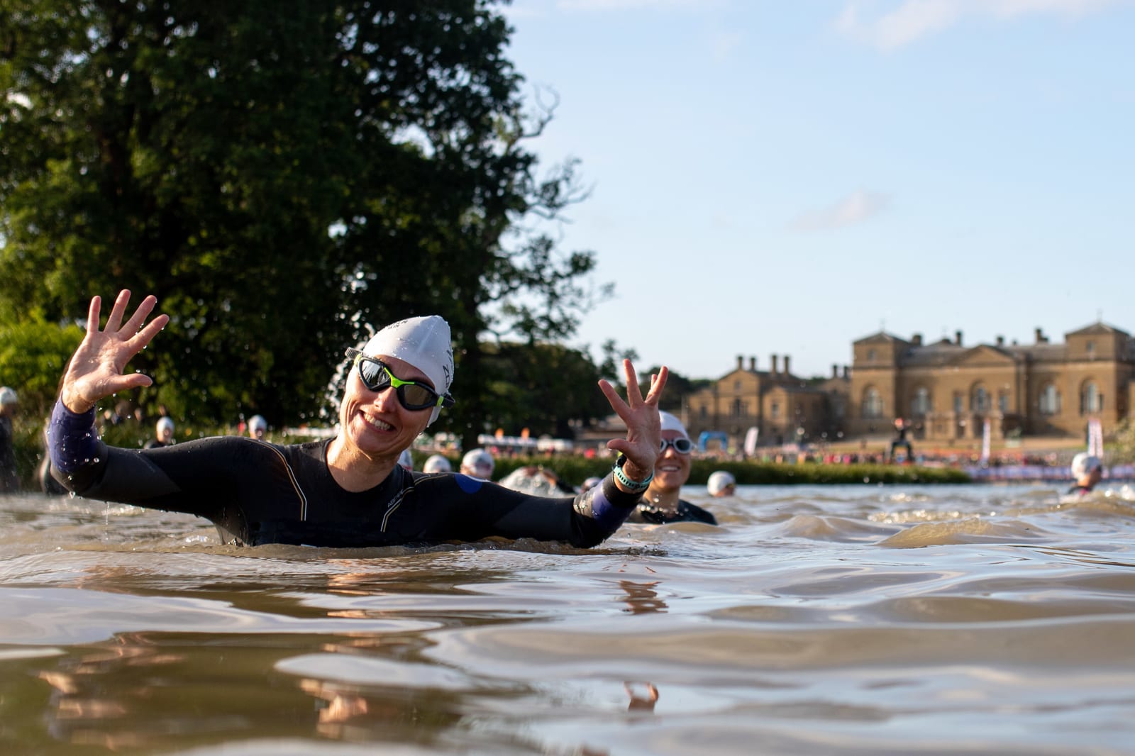 A swimmer wearing a white swim cap and sunglasses smiles and raises both hands while swimming in a body of water. The swimmer is surrounded by others in similar swim attire, with a large building and trees in the background under a clear sky.