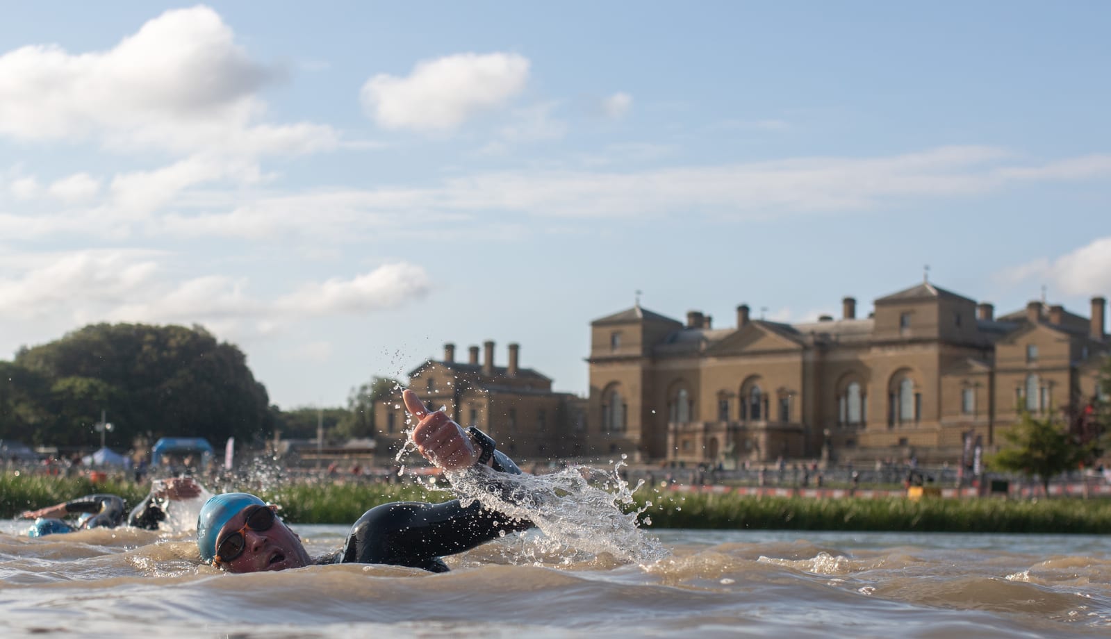 A group of swimmers in wetsuits and swim caps participate in an outdoor swimming event, with one swimmer in the foreground performing a front crawl. The background shows a large, historic building with a clear, sunny sky overhead.