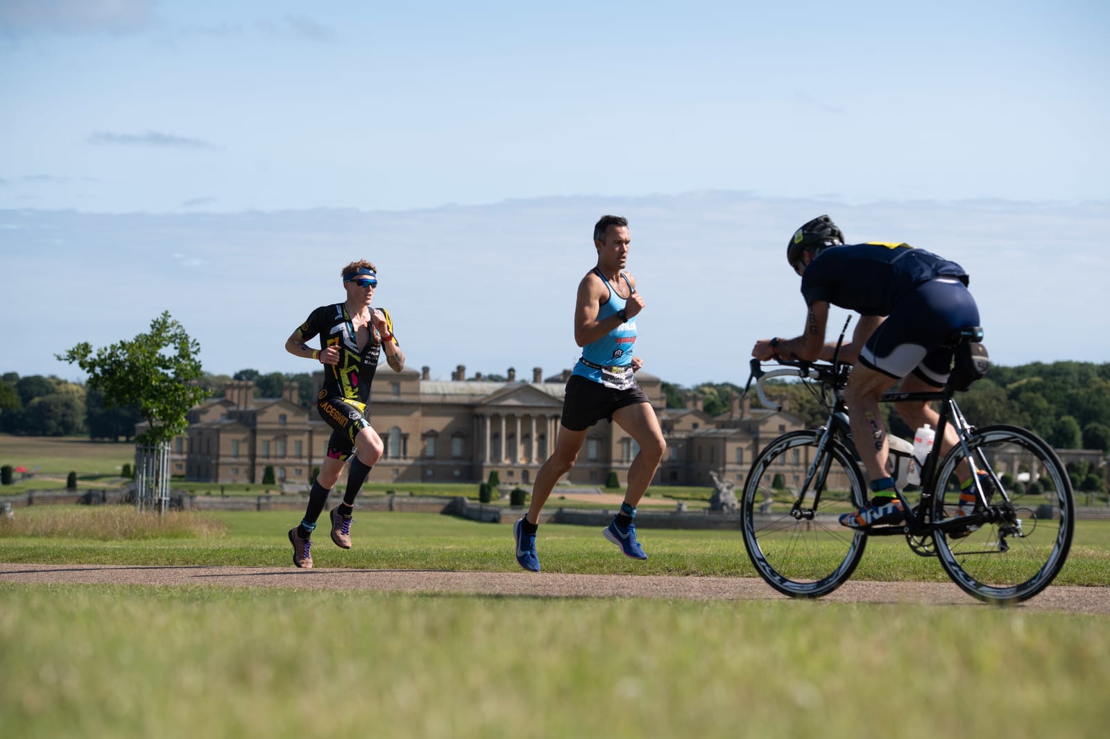 A cyclist and two runners participate in a triathlon event in a scenic outdoor area with an historic building in the background. The sky is clear and the participants appear focused on the race. Grass and trees are visible along the path.