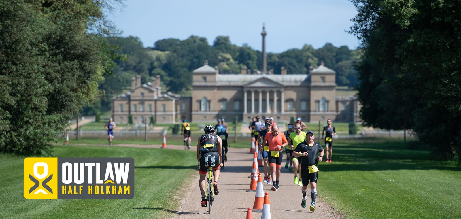 Participants are running and cycling on a path towards a grand historical building during the Outlaw Half Holkham triathlon event. The sun is shining and trees line the path, which is sectioned off with cones. The event logo is visible in the bottom left corner.