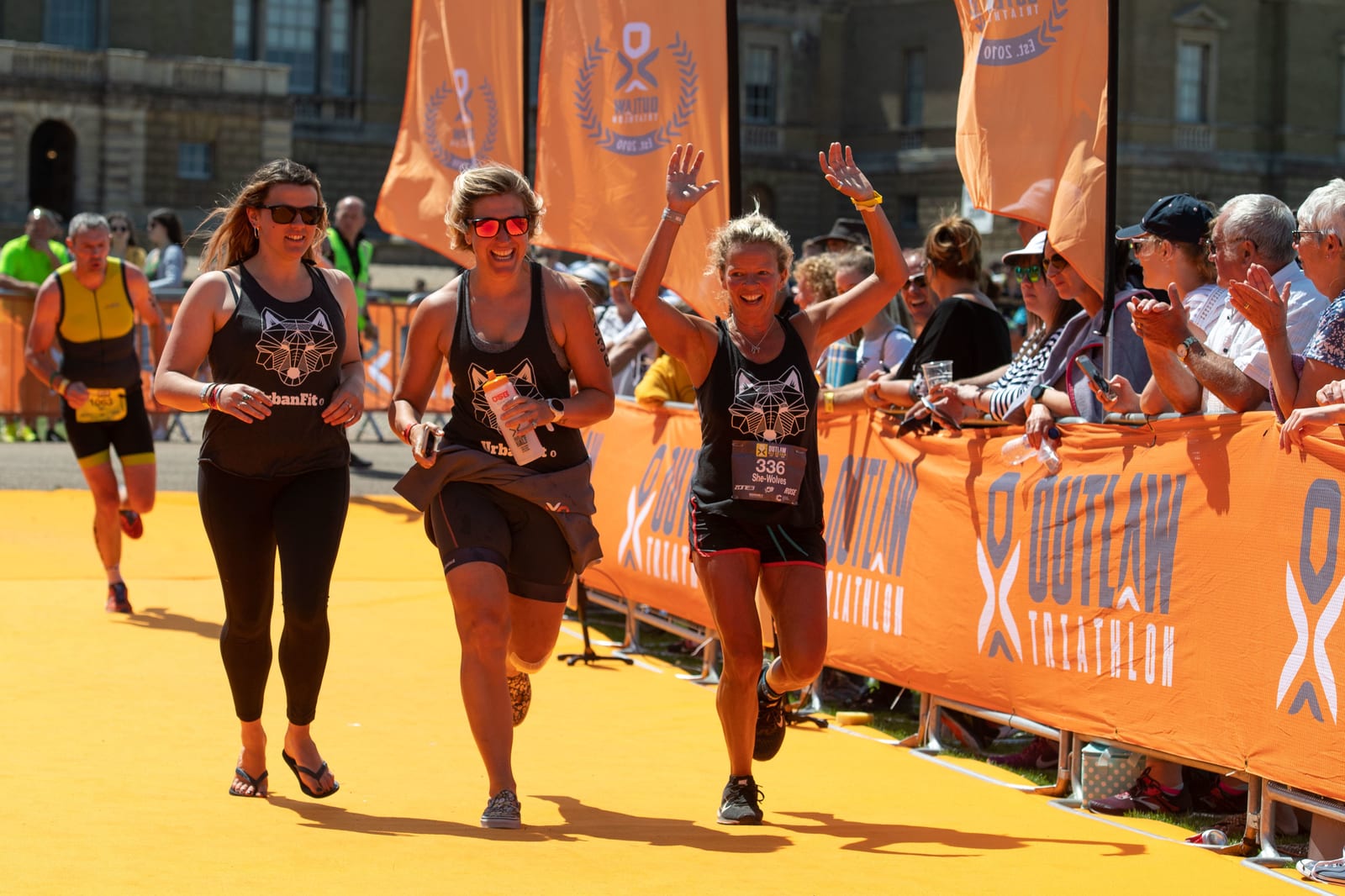 Three jubilant women, wearing athletic gear and race number bibs, run hand-in-hand towards an orange finish line, high-fiving spectators. Behind them, other participants are visible under sunny skies, with an encouraging crowd lining the race pathway.