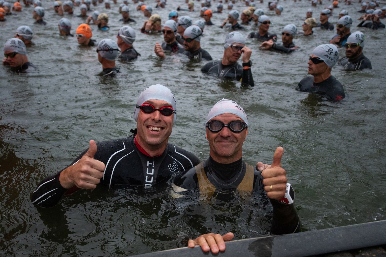 A large group of swimmers in wetsuits and swim caps stand in a lake, preparing for a race. Two men in the foreground give a thumbs up and smile at the camera, while other participants adjust their gear and wait for the event to start.