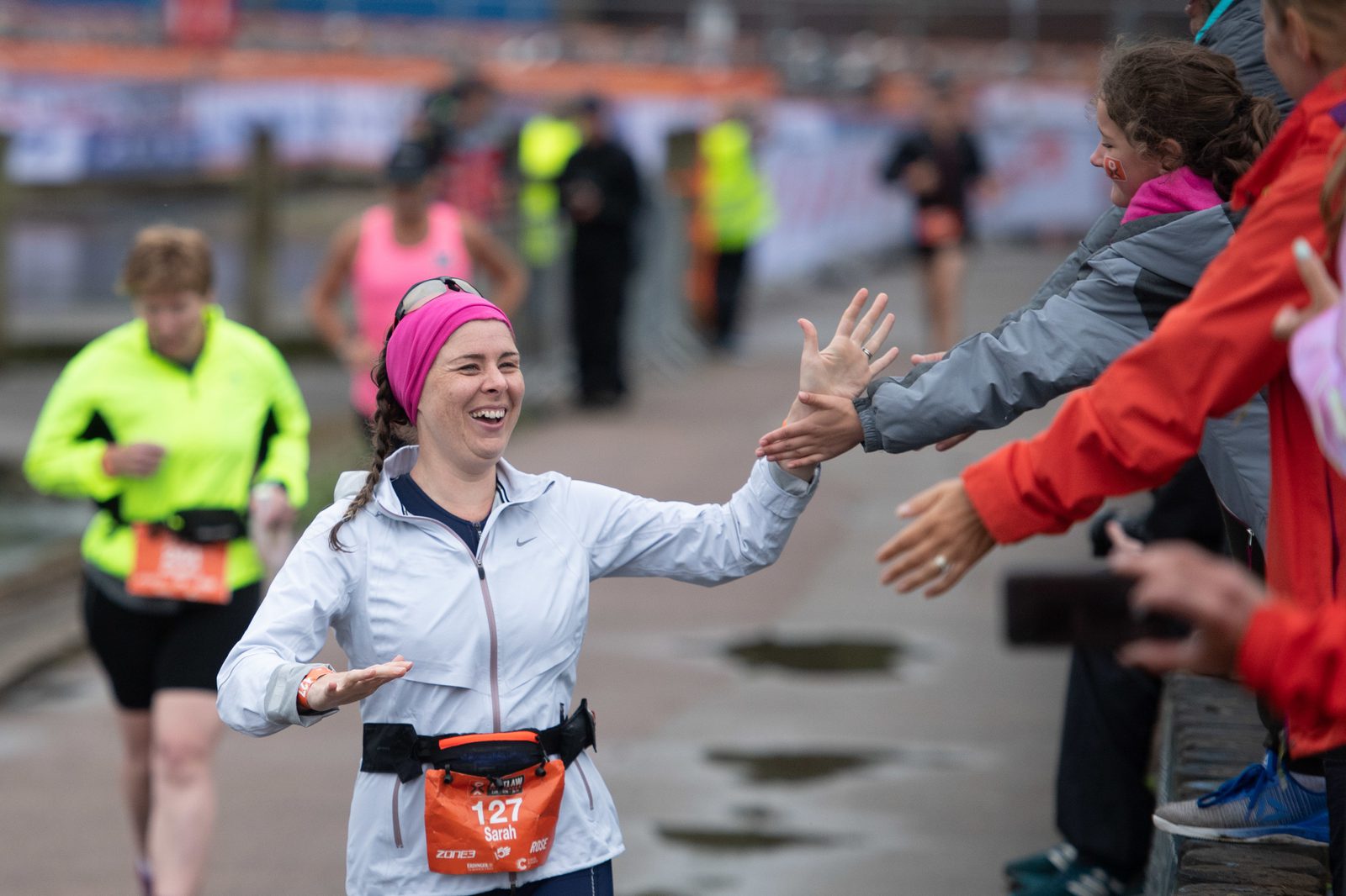 A smiling marathon runner in a white jacket and pink headband high-fives a person from the crowd. She is wearing a race bib numbered 127. Other runners and spectators can be seen in the background, with barriers and event staff along the path.