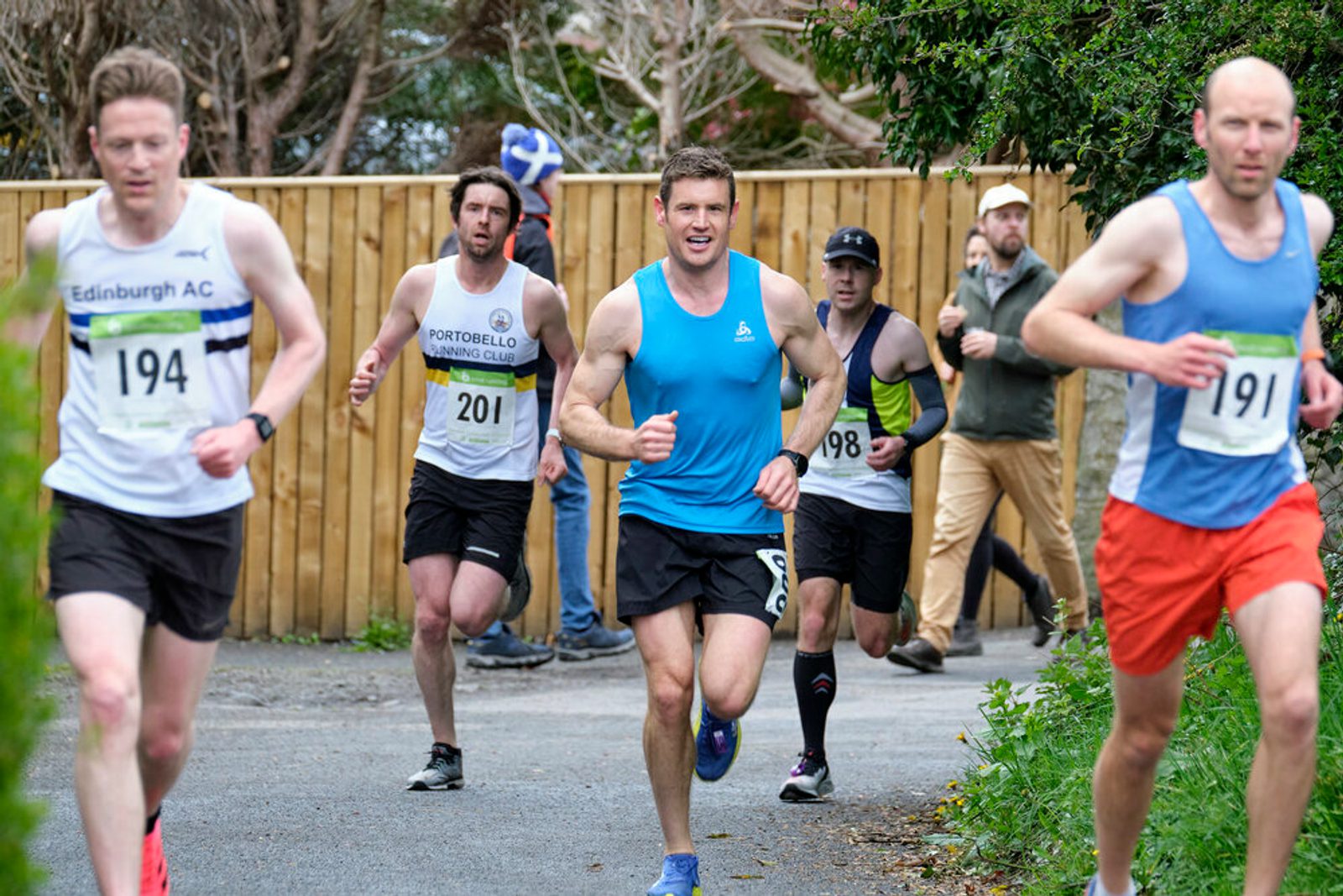 A group of participants, all male and dressed in various athletic gear, are running in a road race. The runner in a blue top and black shorts is in the center, while other runners are visible in the background and foreground. Trees and a wooden fence line the route.