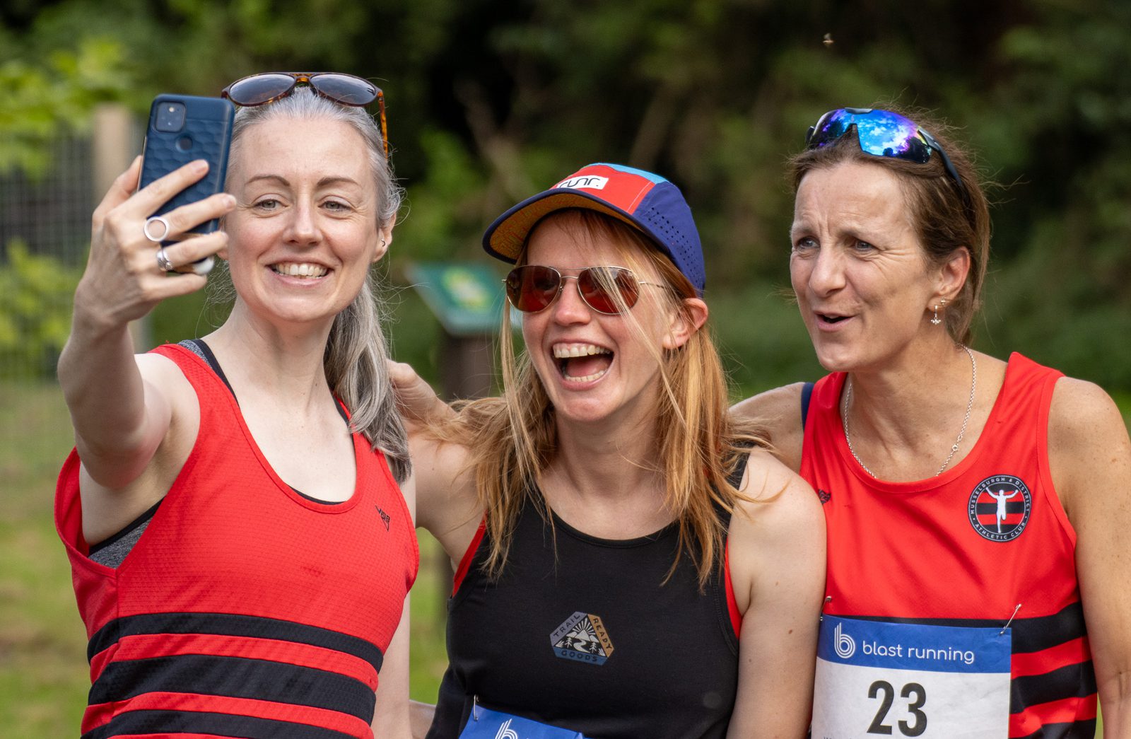 Three women in athletic gear, smiling and posing for a selfie outdoors after a running event. Two wear red running vests and race bibs, while one wears sunglasses and a blue-red cap. Greenery is visible in the background.