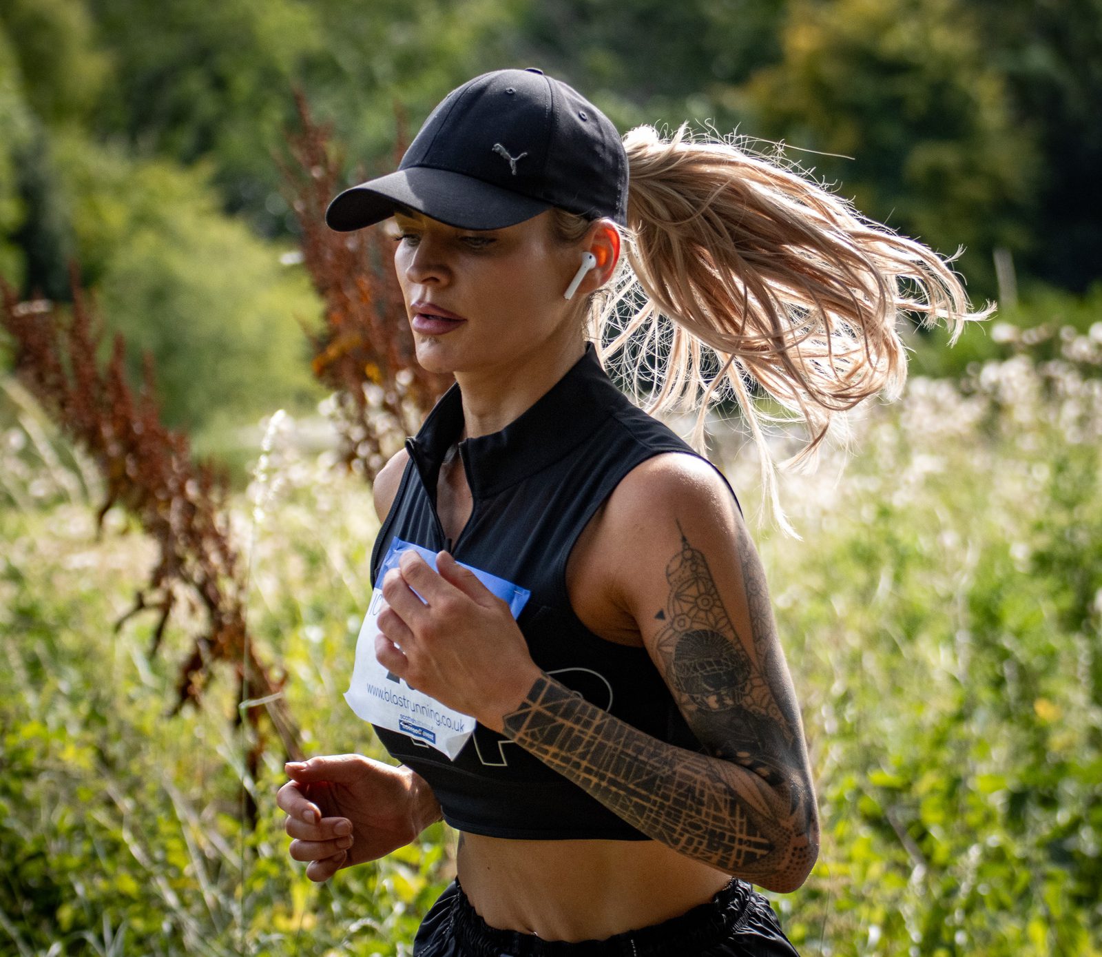 A woman with long blonde hair in a ponytail, wearing a black cap, sleeveless top, and earbuds, runs outdoors through a green, sunlit area. She has a visible arm tattoo and a race bib pinned to her shirt.