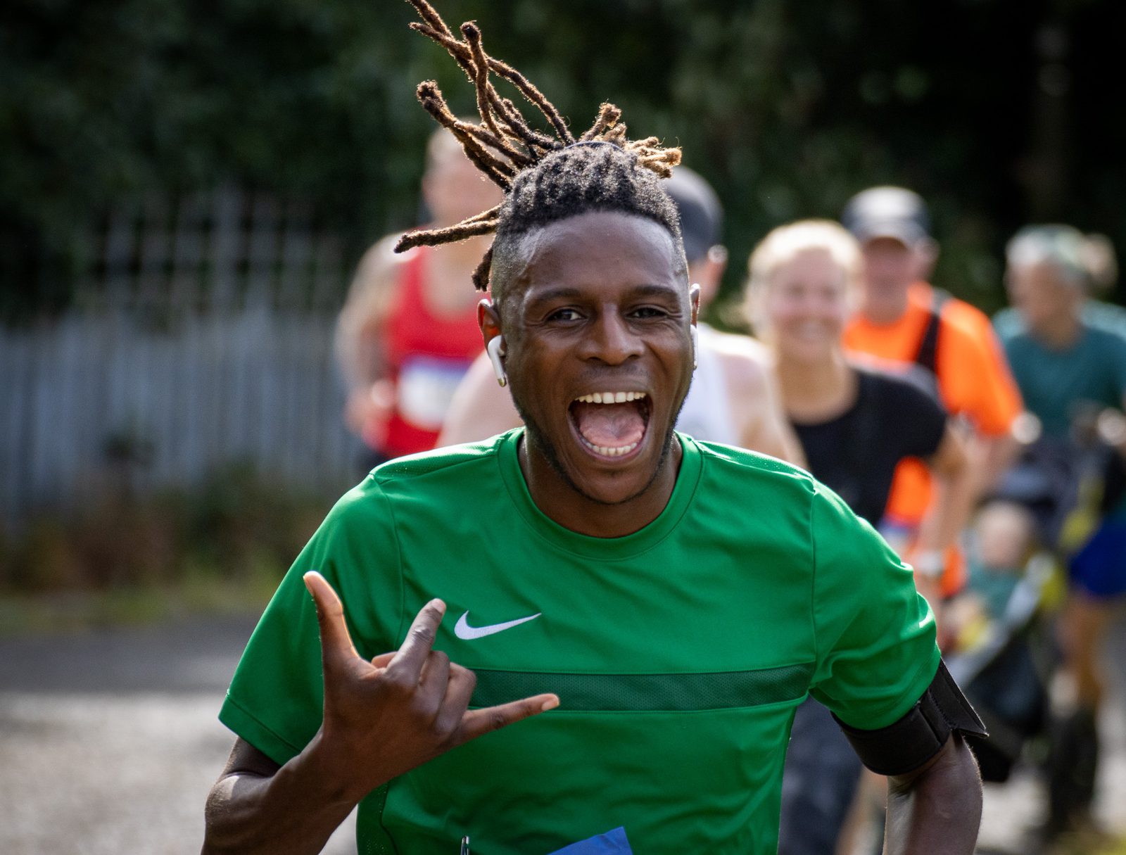 A smiling runner in a green shirt makes a hand gesture while running outdoors, with other runners blurred in the background. The runner has braided hair and looks energetic and happy.