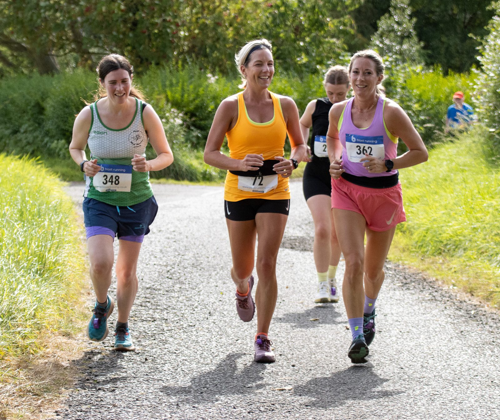 Three women wearing race bibs run together on a sunny path, smiling and chatting. They are dressed in athletic gear, surrounded by green foliage and grass, with another runner visible in the background.