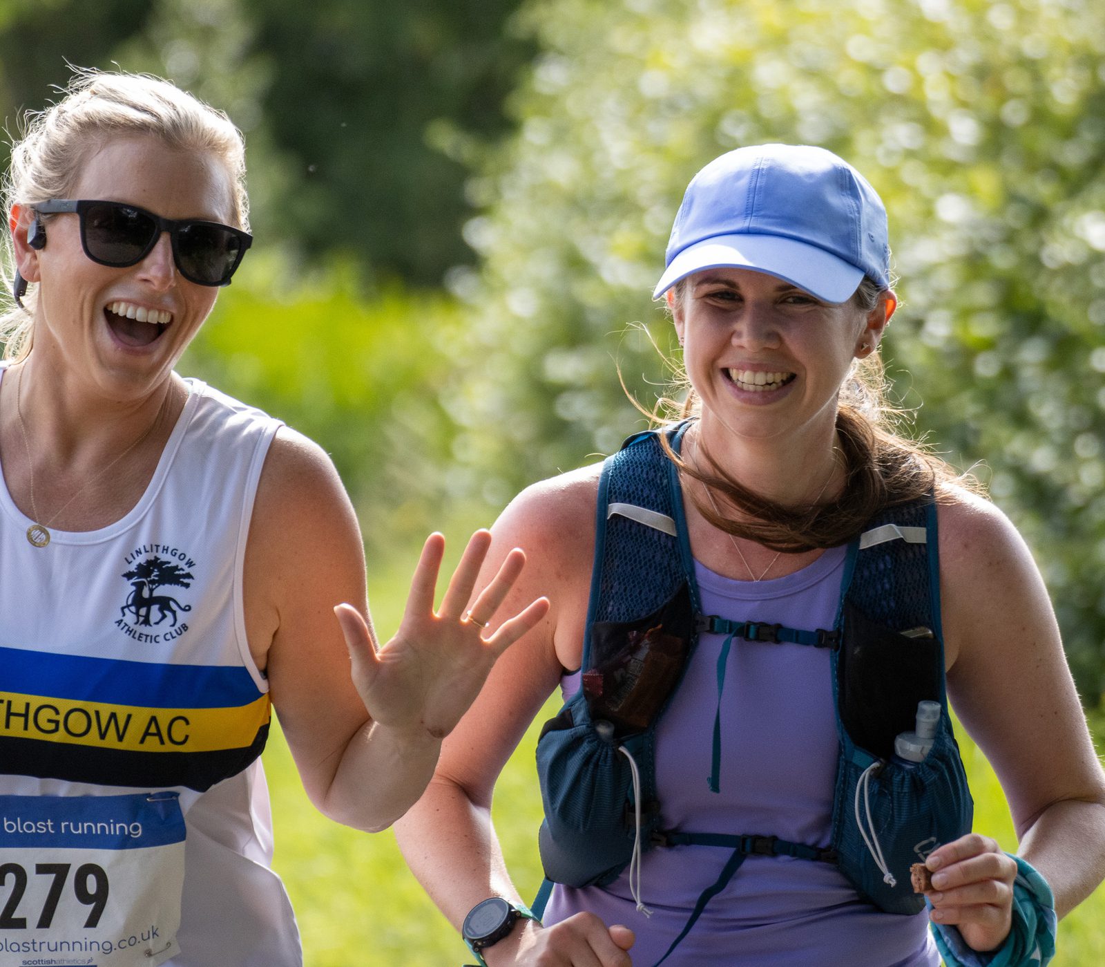Two women smile and wave while running outdoors. One wears sunglasses and a white running vest, the other wears a blue cap and hydration vest. Green foliage is blurred in the background.