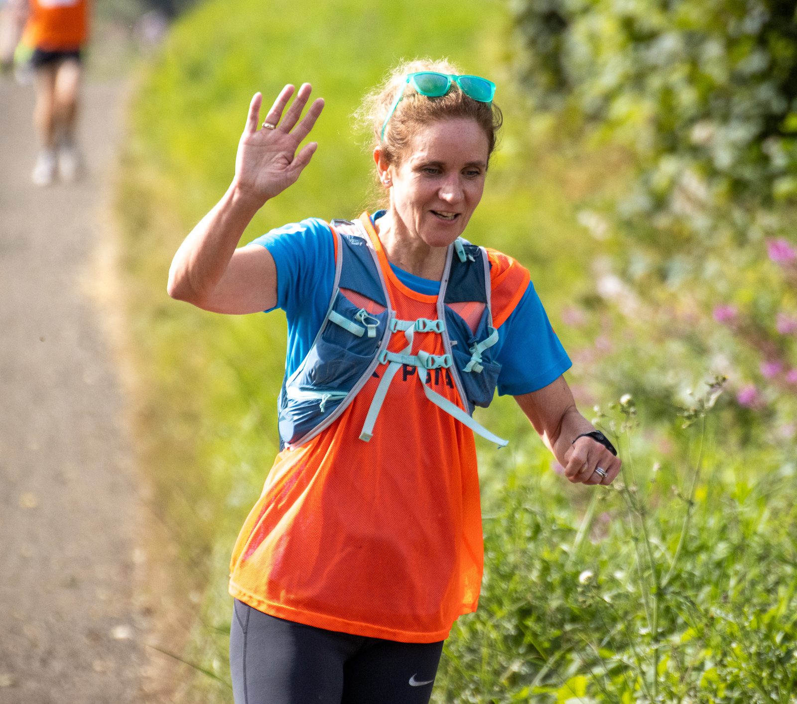 A woman wearing an orange and blue running outfit waves while running outdoors on a sunny day, with greenery and flowers in the background.