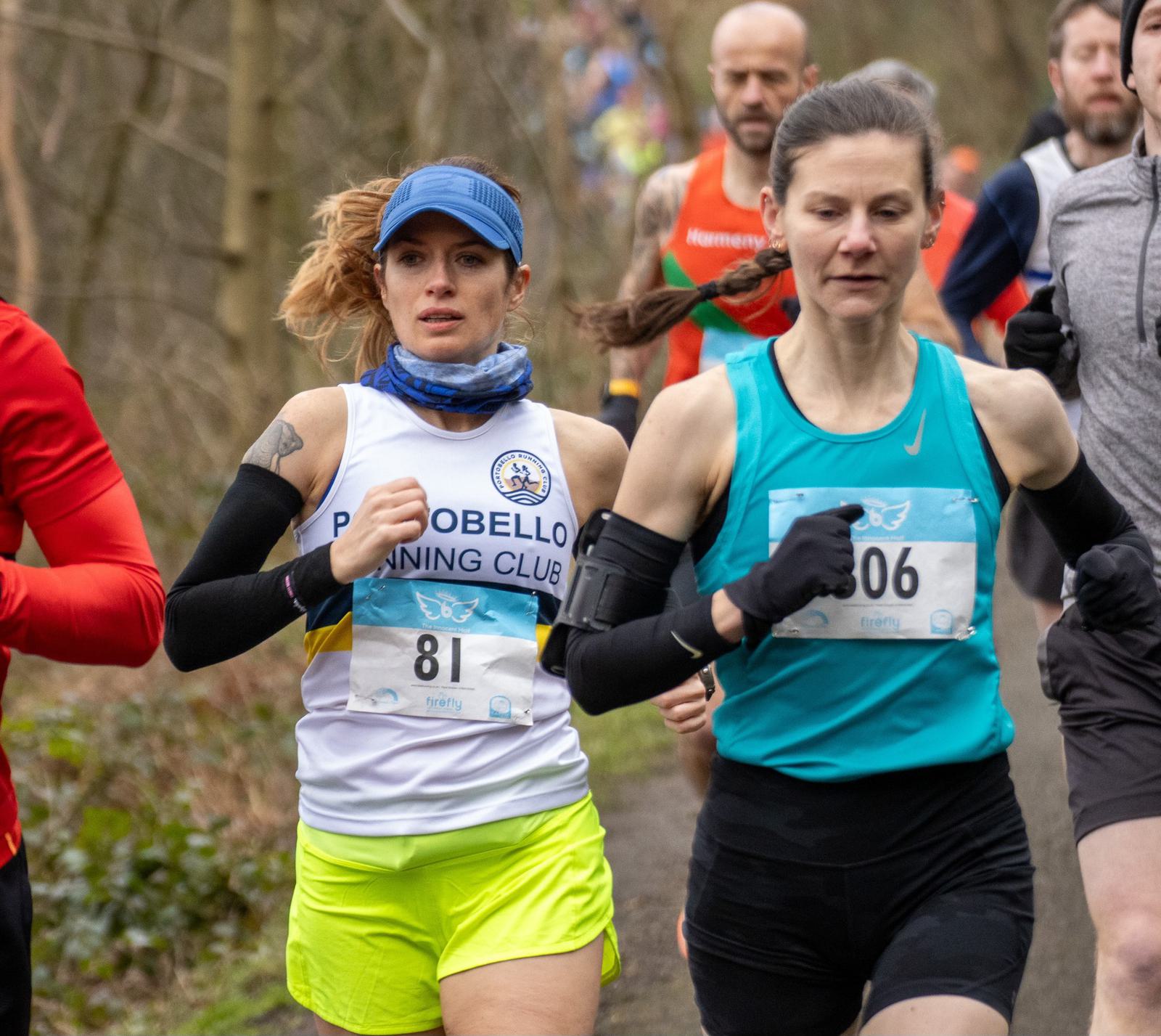 Two runners in a race, one in a white and yellow outfit with a blue cap, and another in a turquoise tank top. Other runners are visible in the background on a wooded path.