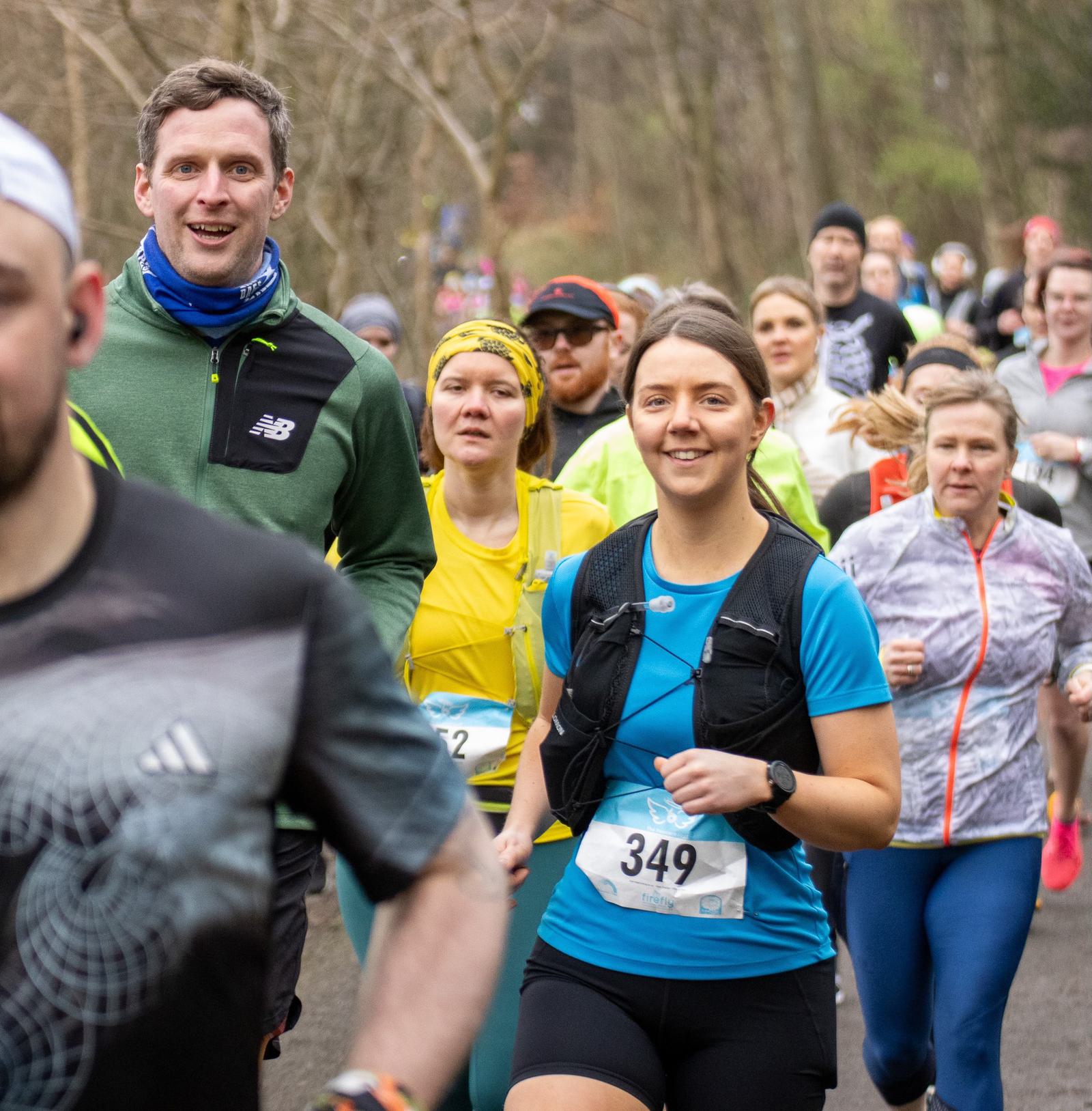 A group of runners participating in a race on a wooded path. The focus is on a man in a green jacket and a woman in a blue shirt with a race bib numbered 349. They are surrounded by other participants in various athletic gear.