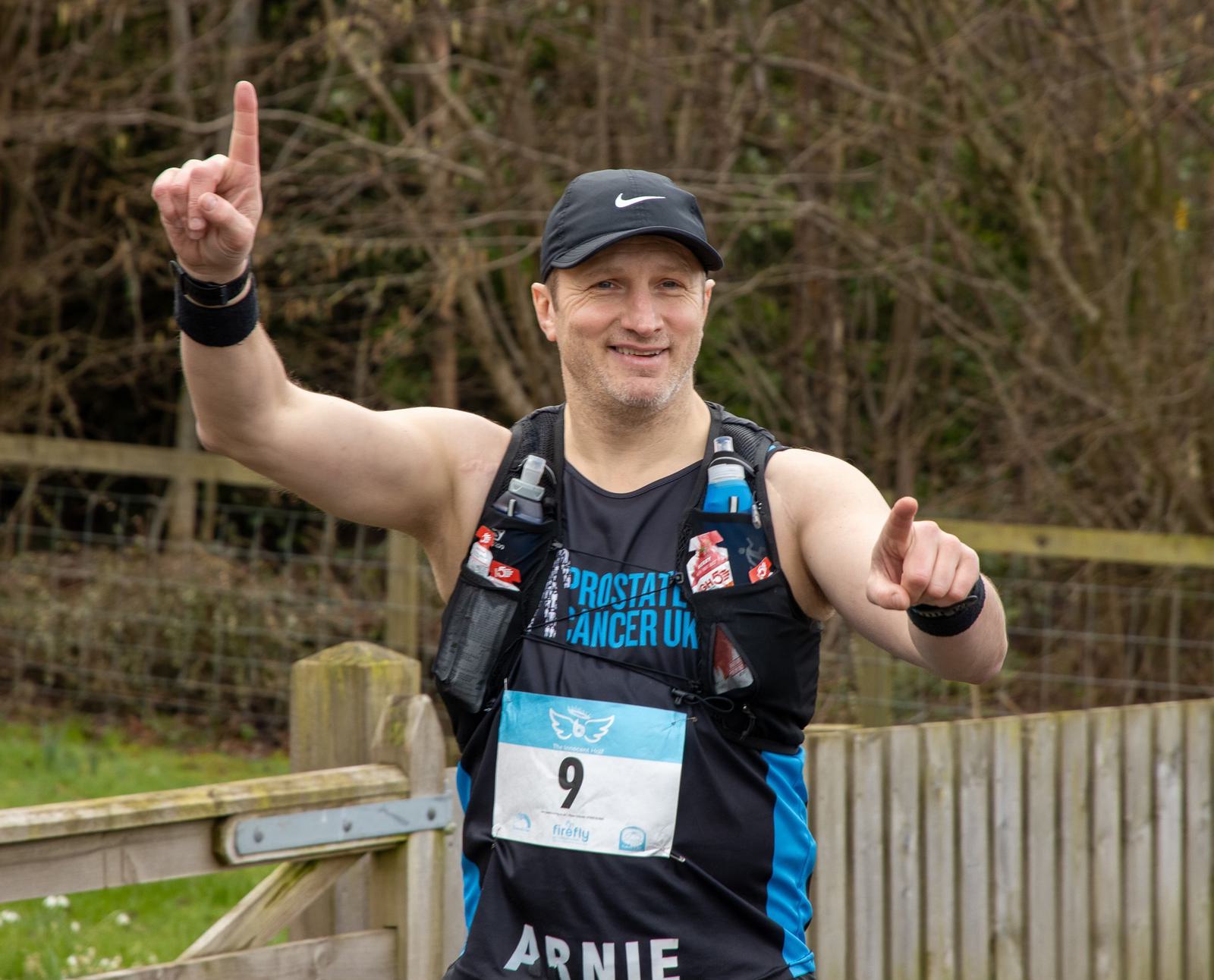 Man in athletic wear and a cap, with a bib number 9, smiles and gestures with both index fingers pointing up. He wears a vest with "Prostate Cancer UK" printed on it. A wooden fence and leafless trees are in the background.