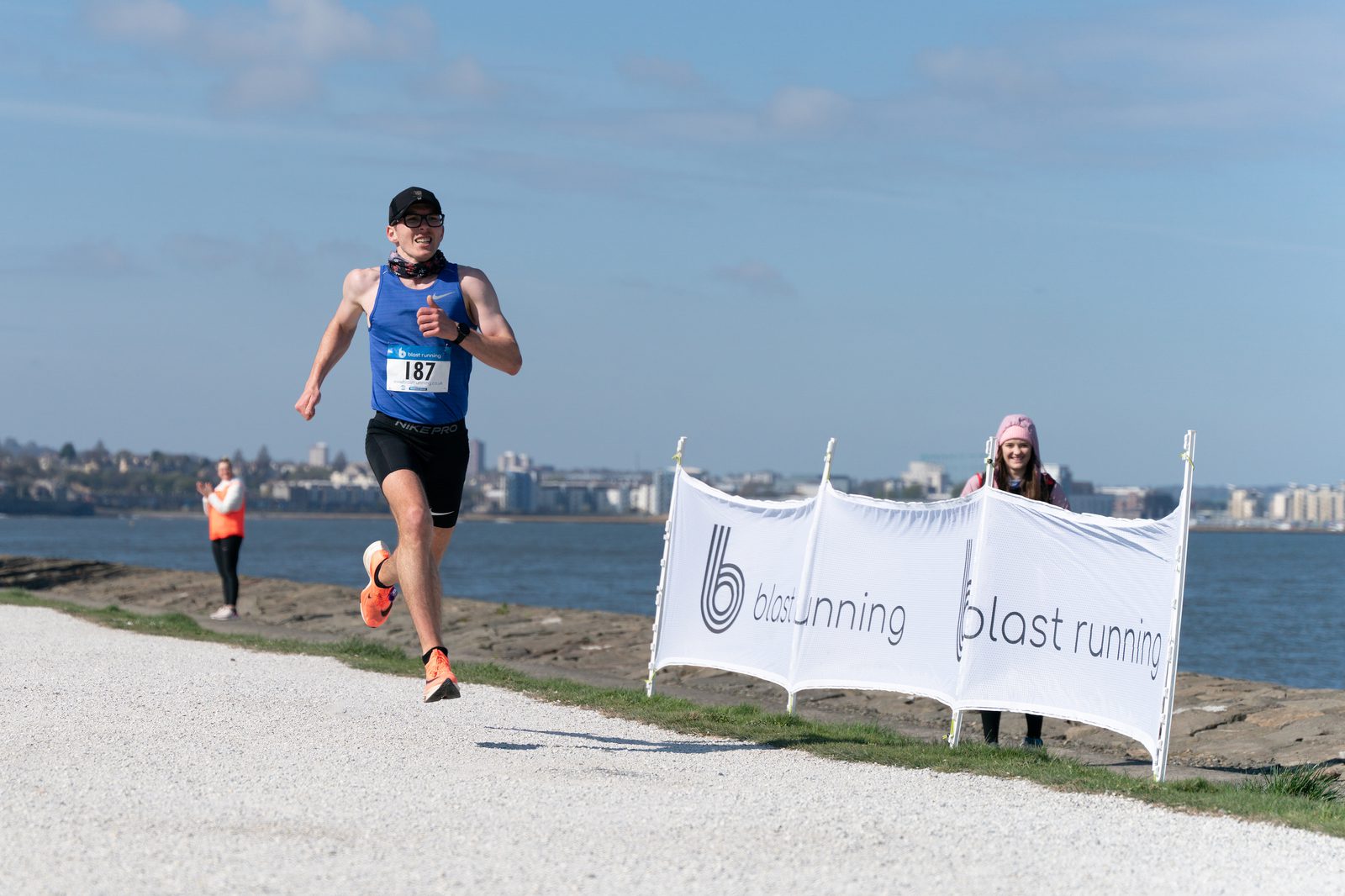 A runner in a blue tank top and black shorts sprints on a gravel path near the waterfront, with a city skyline in the background. He is wearing sunglasses and a cap. Two banners on the right display "blast running," and onlookers in bright vests stand and watch.