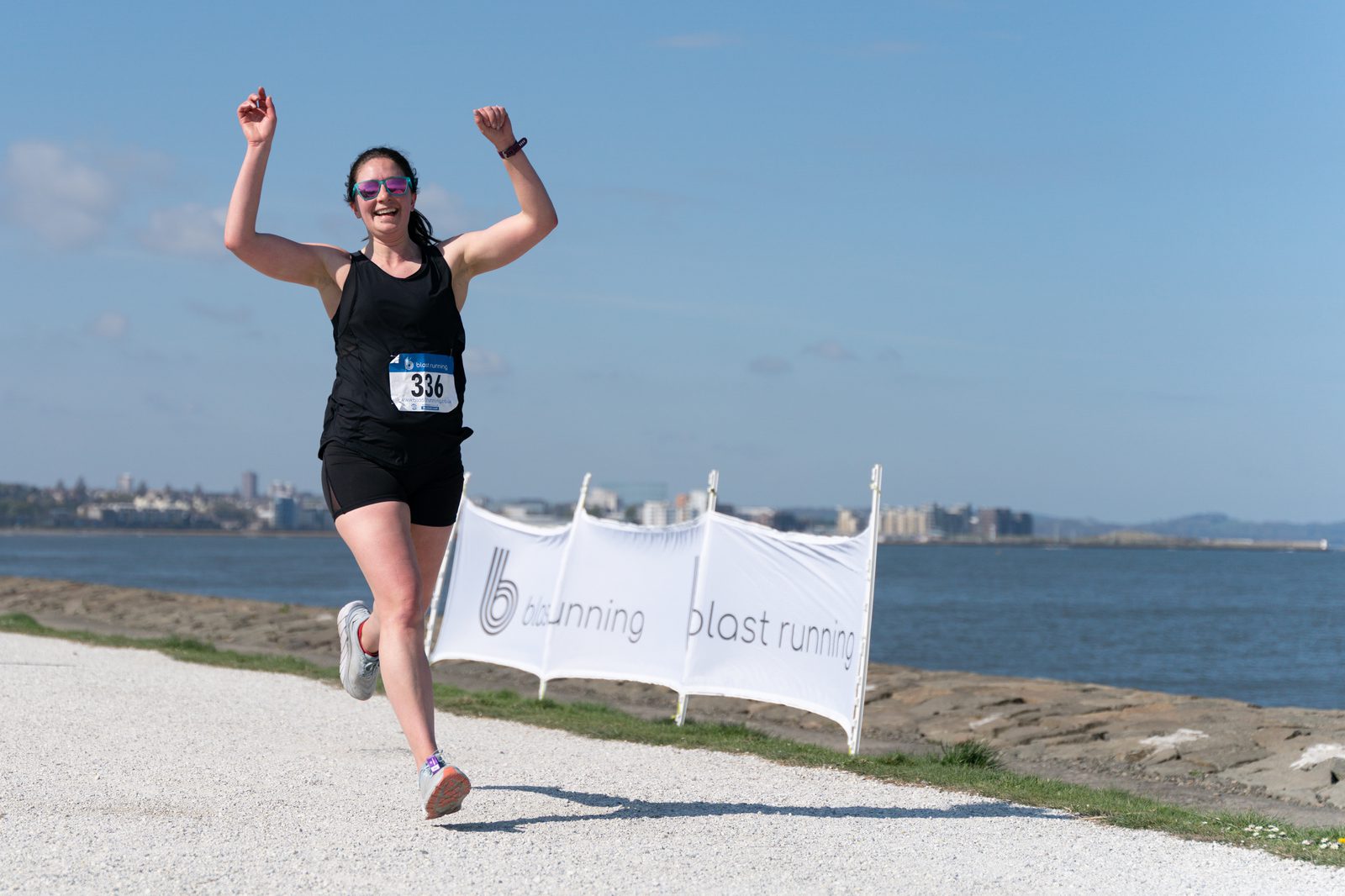 A person in a black athletic outfit and sunglasses is joyfully running with arms raised along a waterfront path. They have a race number pinned to their chest. A banner that reads "blast running" is visible in the background.