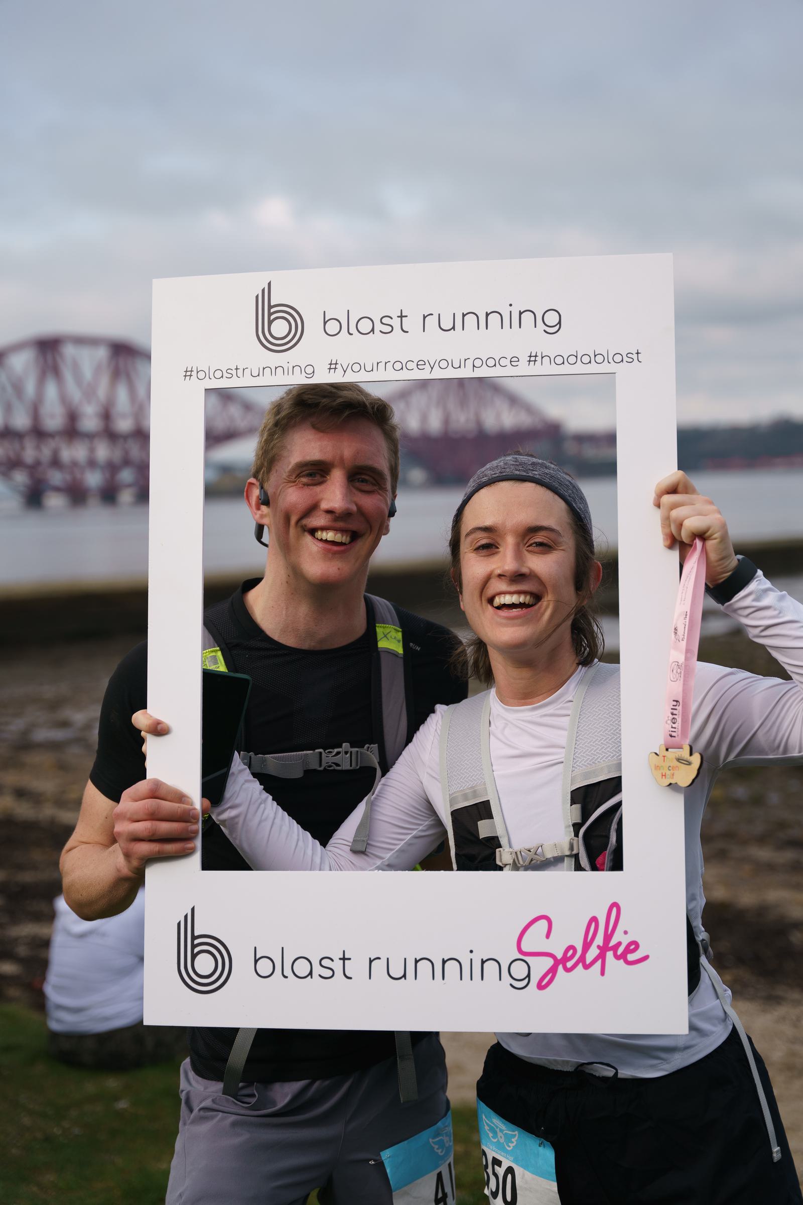 Two people smiling, holding a white frame with "blast running" and hashtags. They're outdoors, wearing athletic gear, with race bibs. One holds a medal. A bridge and water are visible in the background.