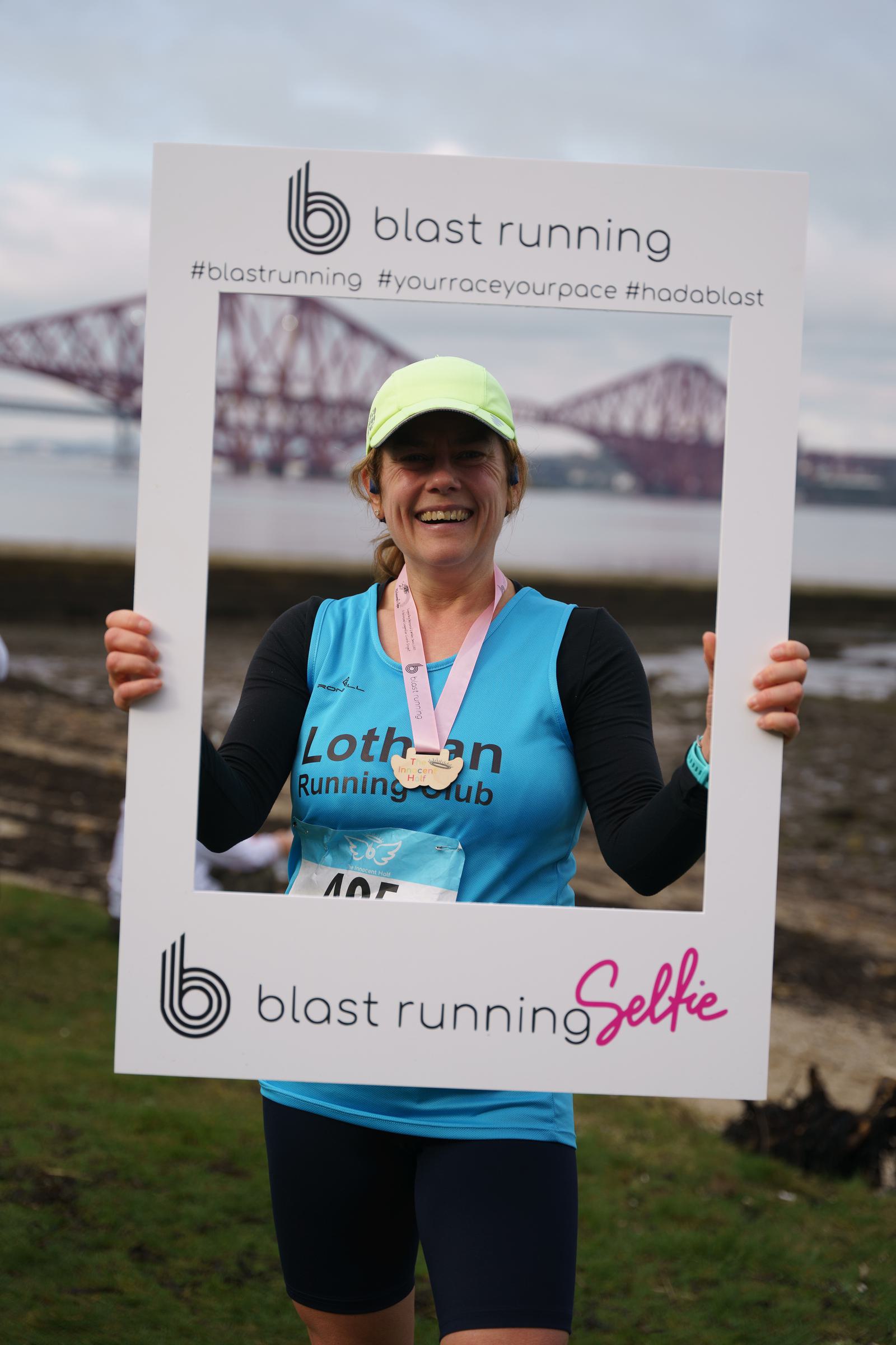A smiling runner wearing a blue "Lothian Running Club" shirt and a medal poses with a "Blast Running" frame near a scenic waterfront, with a large bridge in the background.