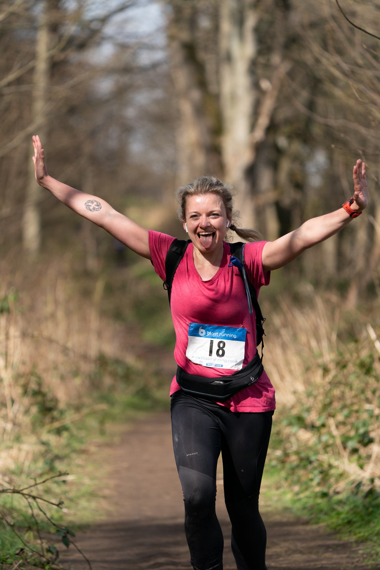A woman in a pink shirt, black leggings, and a sports watch, with a race number "18" on her front, runs on a wooded trail. She is smiling, sticking her tongue out, and has her arms raised in excitement. The background shows a blurred pathway and trees.